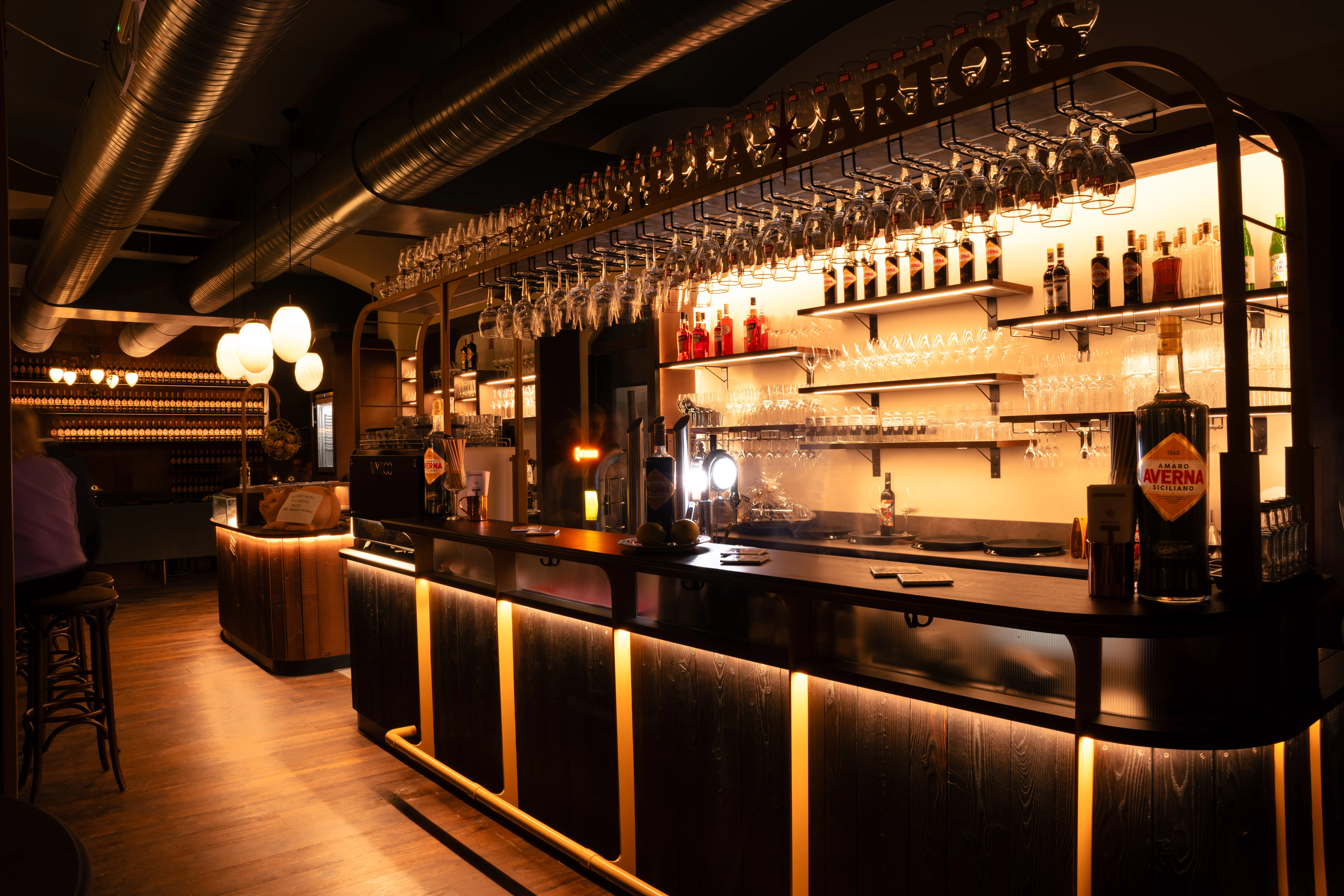 Stilvolle Bar mit dunklem Holz, beleuchtet mit warmweißem Licht. Viele Gläser und Flaschen mit Spirituosen sind zu sehen. Foto von Simon Elias Leser.