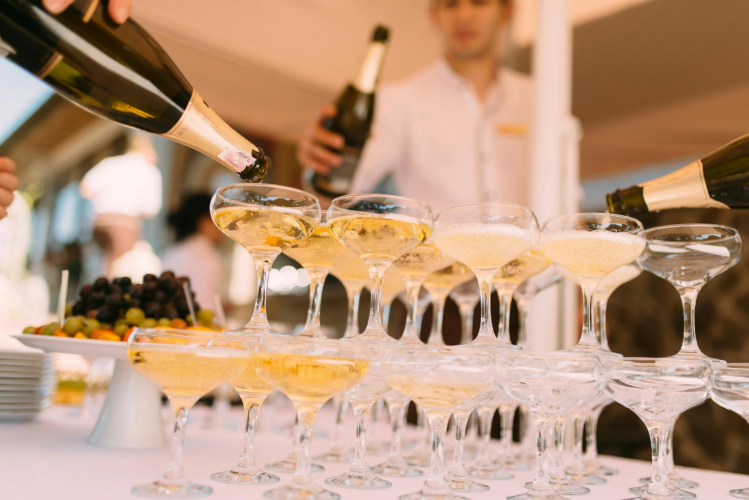 Das Bild zeigt einen festlichen Champagnerturm, in den mehrere Personen gleichzeitig Sekt eingießen. Die Gläser sind elegant gestapelt, der Schaumwein fließt von oben nach unten. Im Hintergrund steht Servicepersonal, daneben eine Obstplatte – die Szene wirkt edel und feierlich.