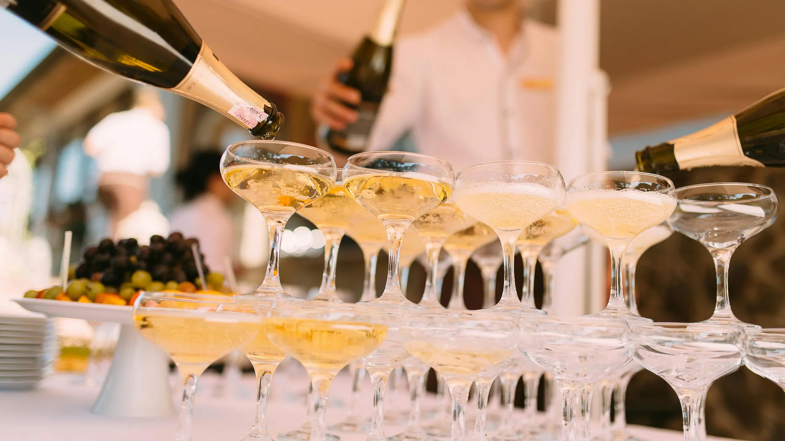 Das Bild zeigt einen festlichen Champagnerturm, in den mehrere Personen gleichzeitig Sekt eingießen. Die Gläser sind elegant gestapelt, der Schaumwein fließt von oben nach unten. Im Hintergrund steht Servicepersonal, daneben eine Obstplatte – die Szene wirkt edel und feierlich.