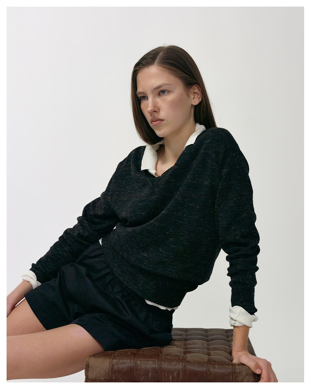 Model mit schwarzem Pullover, weißem Hemd und schwarzen Shorts sitzt vor weißem Hintergrund