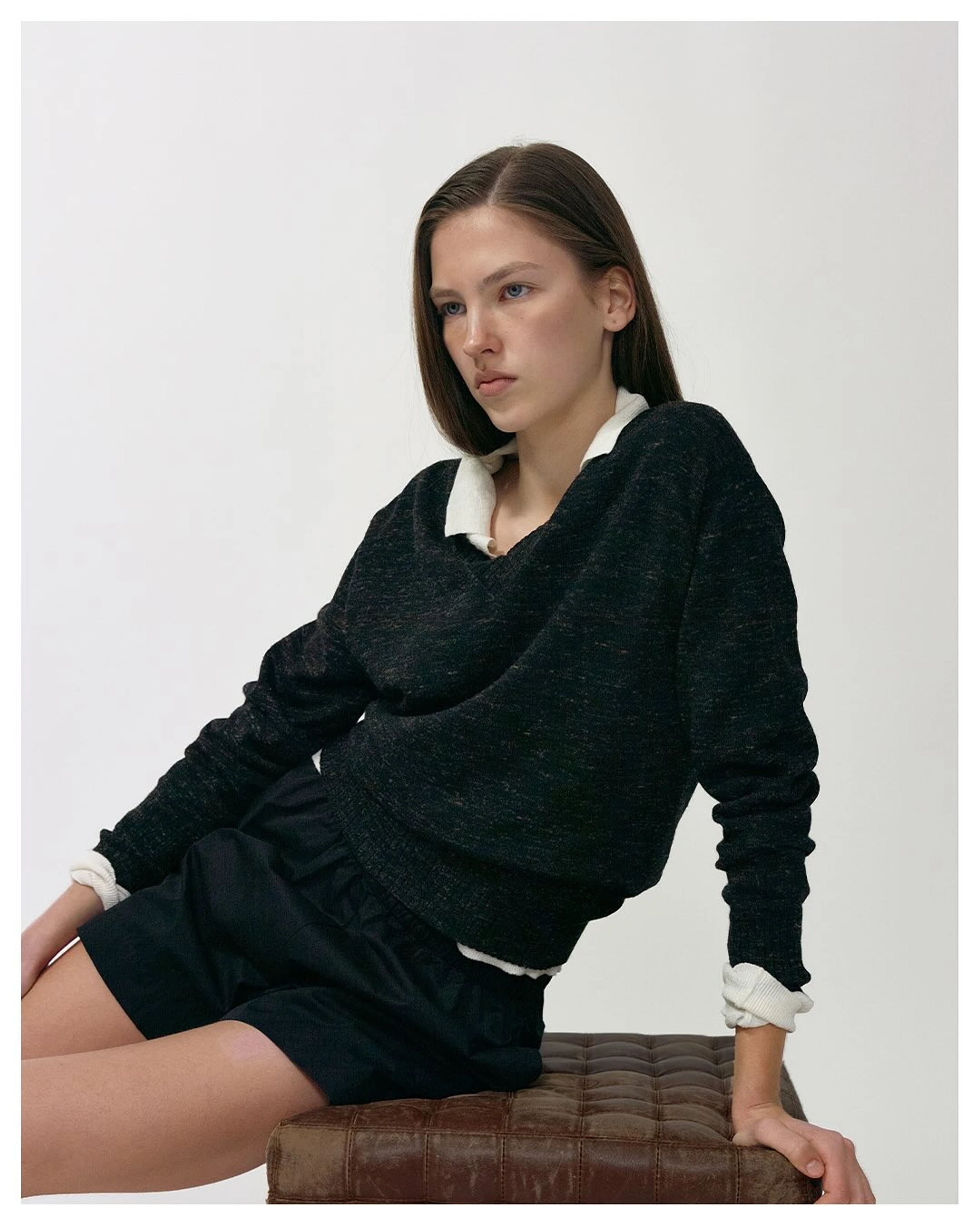 Model mit schwarzem Pullover, weißem Hemd und schwarzen Shorts sitzt vor weißem Hintergrund