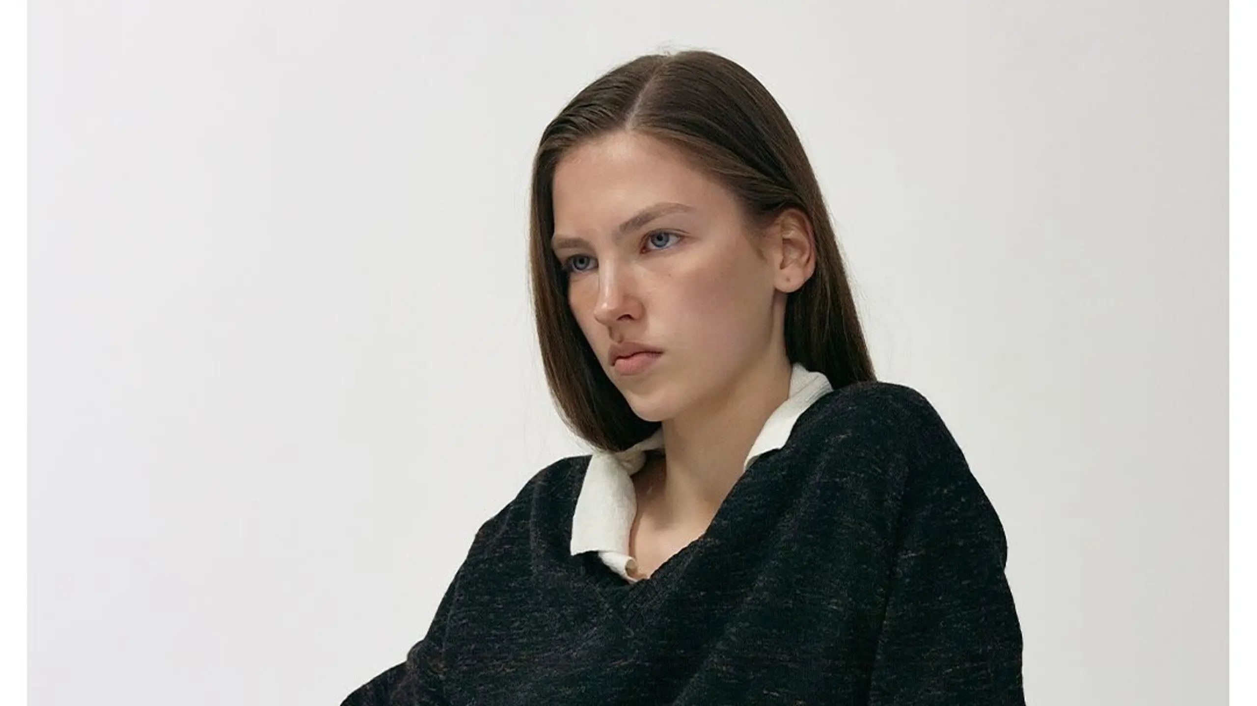Model mit schwarzem Pullover, weißem Hemd und schwarzen Shorts sitzt vor weißem Hintergrund