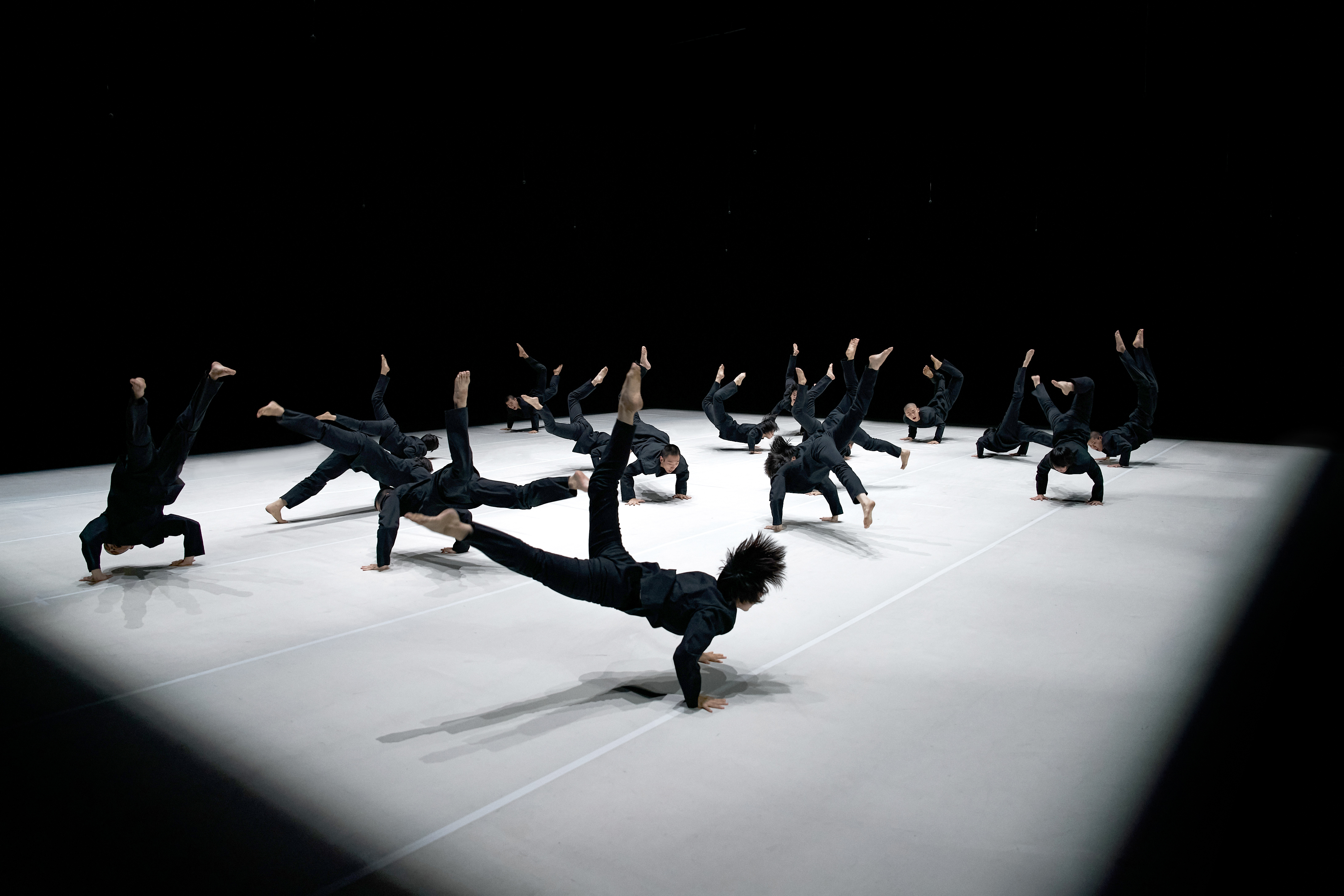 © Fan Xi Moderne Tanzperformance: Mehrere Tänzer in Schwarz führen dynamische Bewegungen auf einer hellen Bühne aus.
