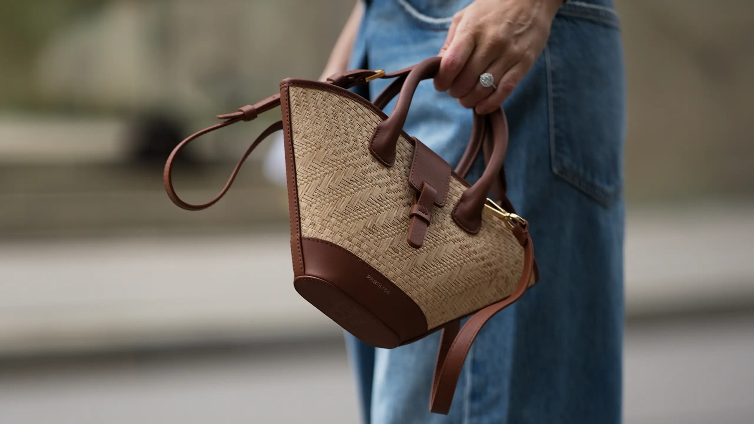 Eine Frau von der Hüfte abwärts. Sie trägt eine blaue Jeans und hält eine Tasche aus Bast in der Hand