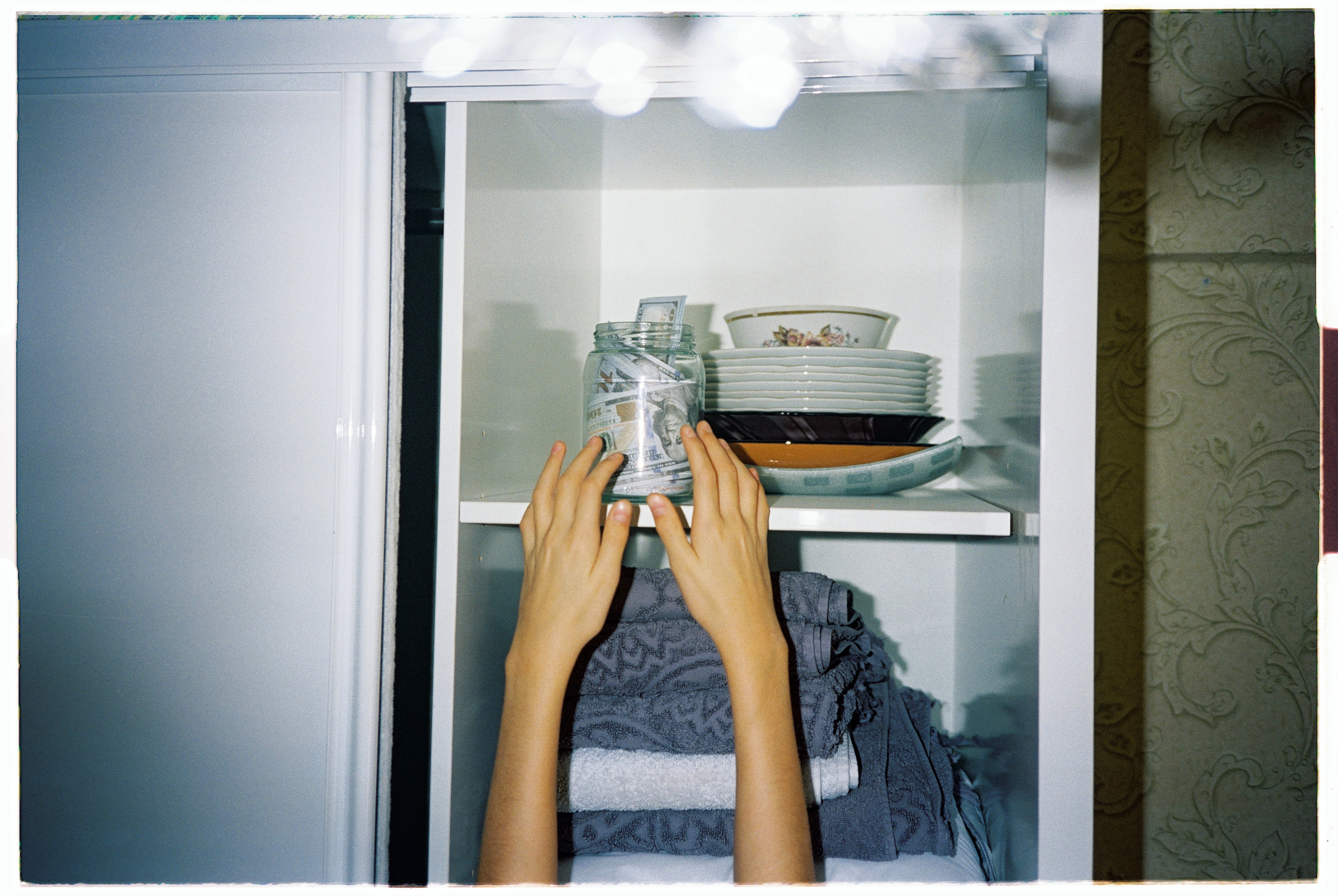 Ein Glas mit Geldscheinen steht oben in einem Regal, zwei Arme strecken