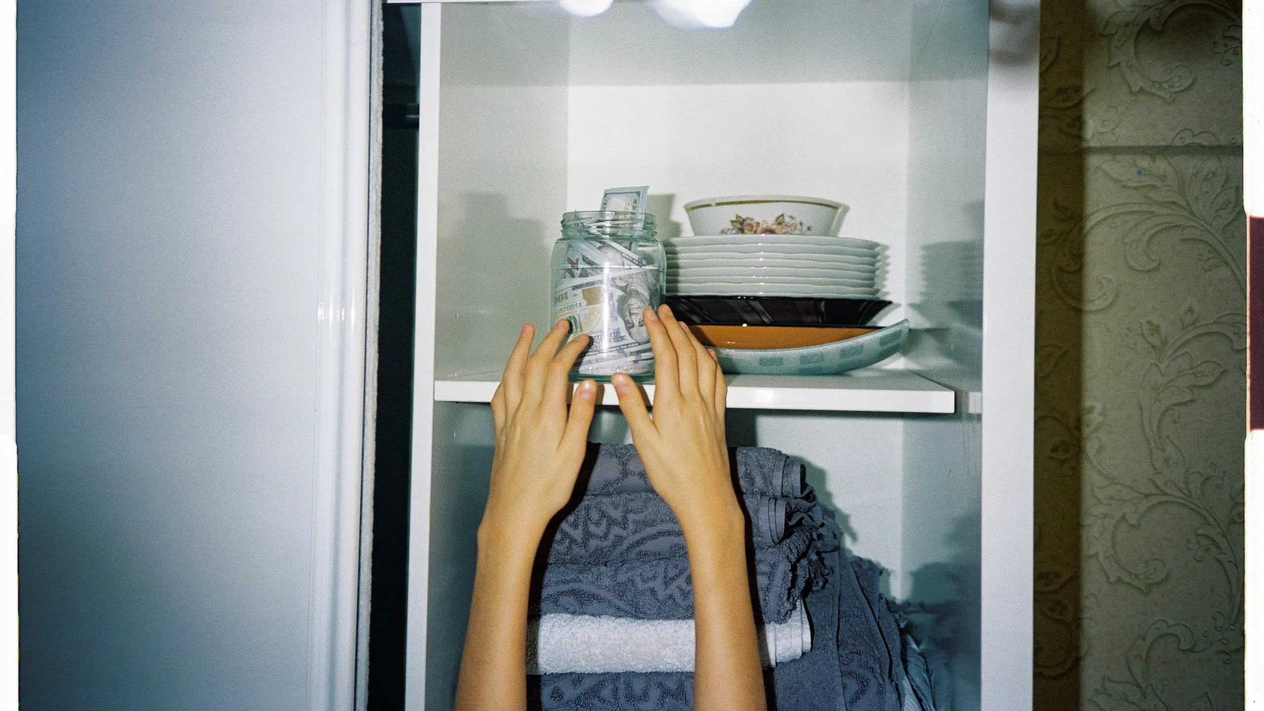 Ein Glas mit Geldscheinen steht oben in einem Regal, zwei Arme strecken