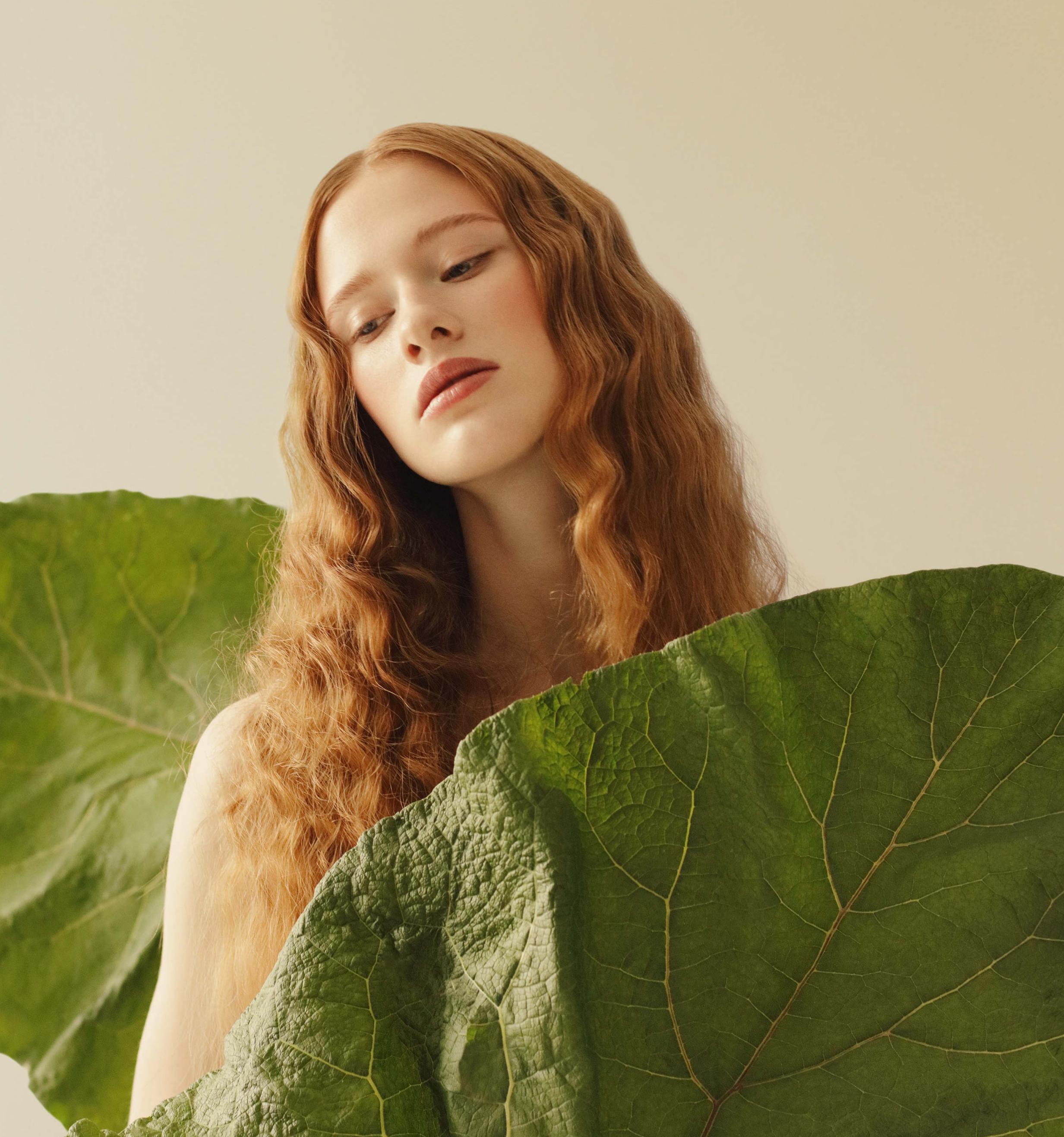 Natürliches Model vor beigem Hintergrund und mit großen grünen Blättern und