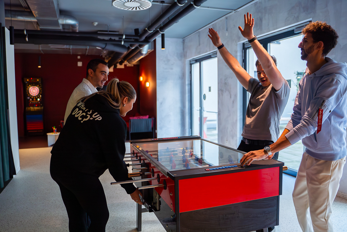 © Vier junge Erwachsene spielen Tischfußball in einem modernen Raum. Spaß und Teamwork im Mittelpunkt.