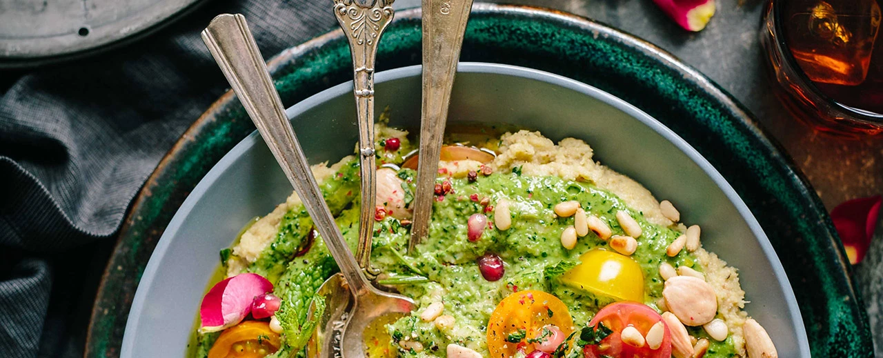 Nahaufnahme einer Schüssel mit einem leckeren, gesunden Gericht aus cremiger Basis, grünem Pesto, Pinienkernen, Mandeln, Tomaten und Granatapfelkernen. Drei antike Löffel liegen in der Schüssel.