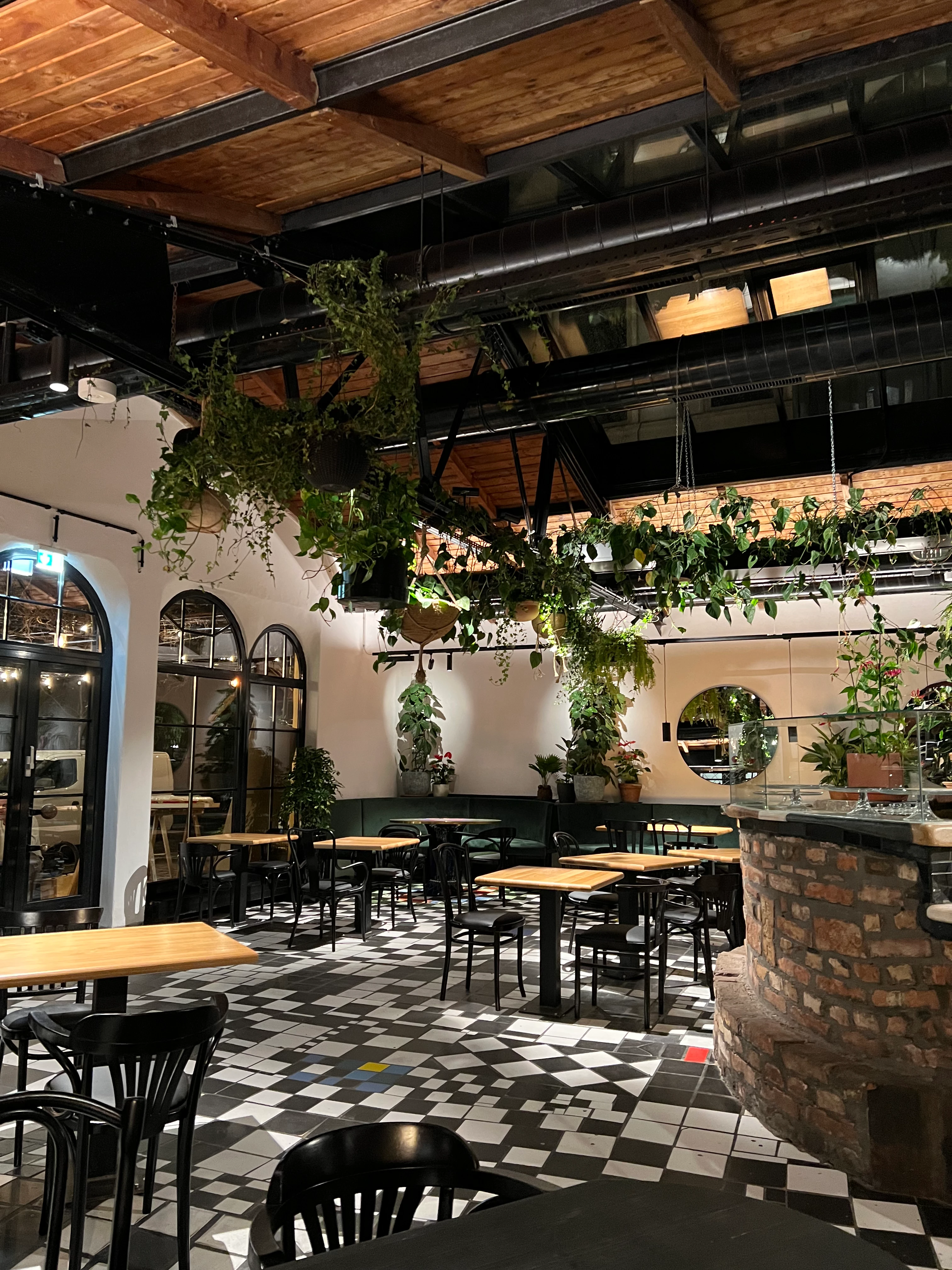 Modernes Restaurant-Interieur mit Pflanzen, Holzbalken & schwarz-weißem Fliesenboden.