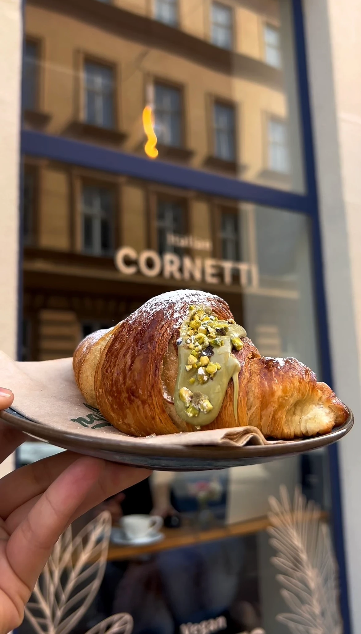 Appetitliches Pistazien-Croissant mit Pistaziencreme und -stückchen, vor einem Café mit der Aufschrift 