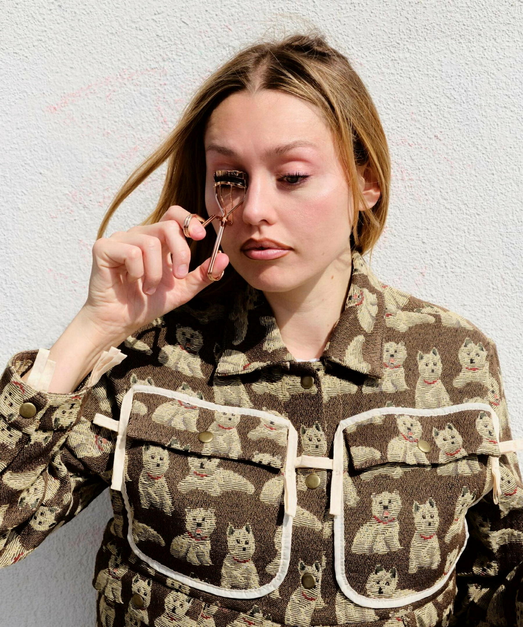 Emily Wood wie sie mit einer WImpernzange ihre WImpern beigt, draußen vor weißer Hauswand