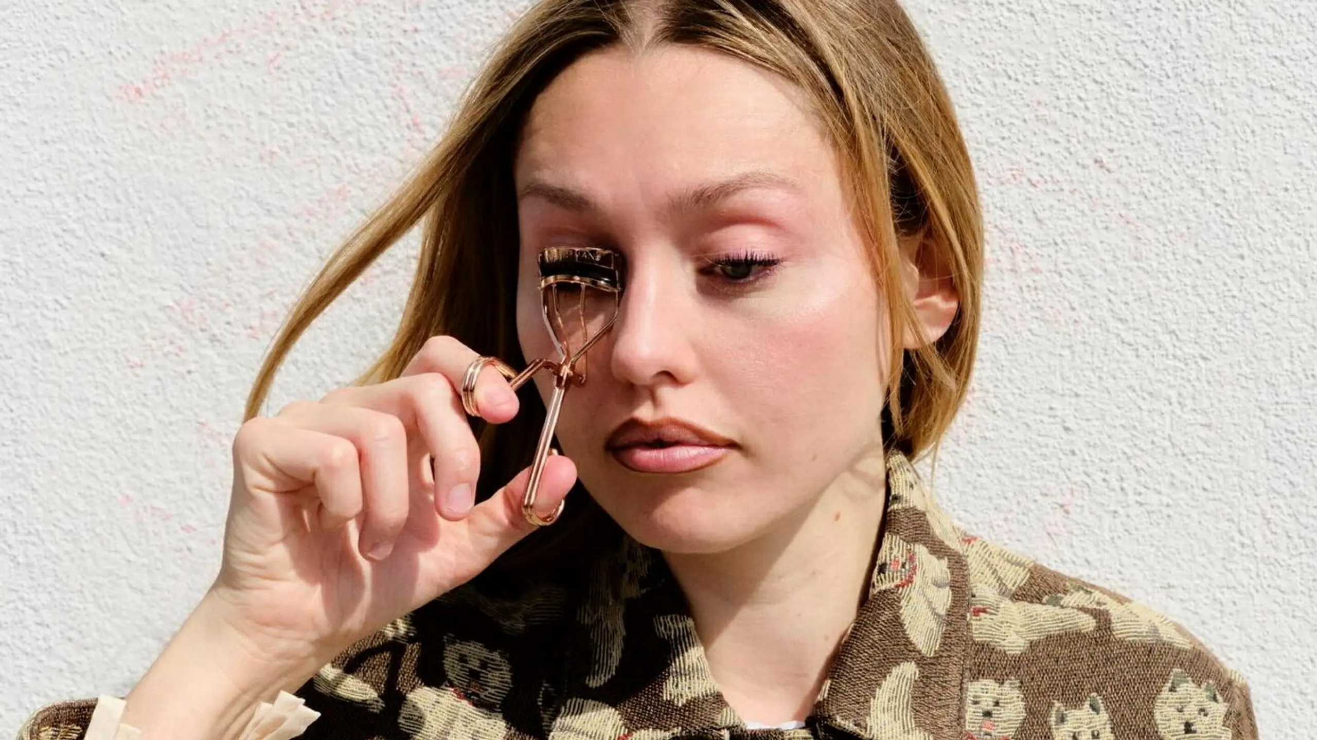 Emily Wood wie sie mit einer WImpernzange ihre WImpern beigt, draußen vor weißer Hauswand