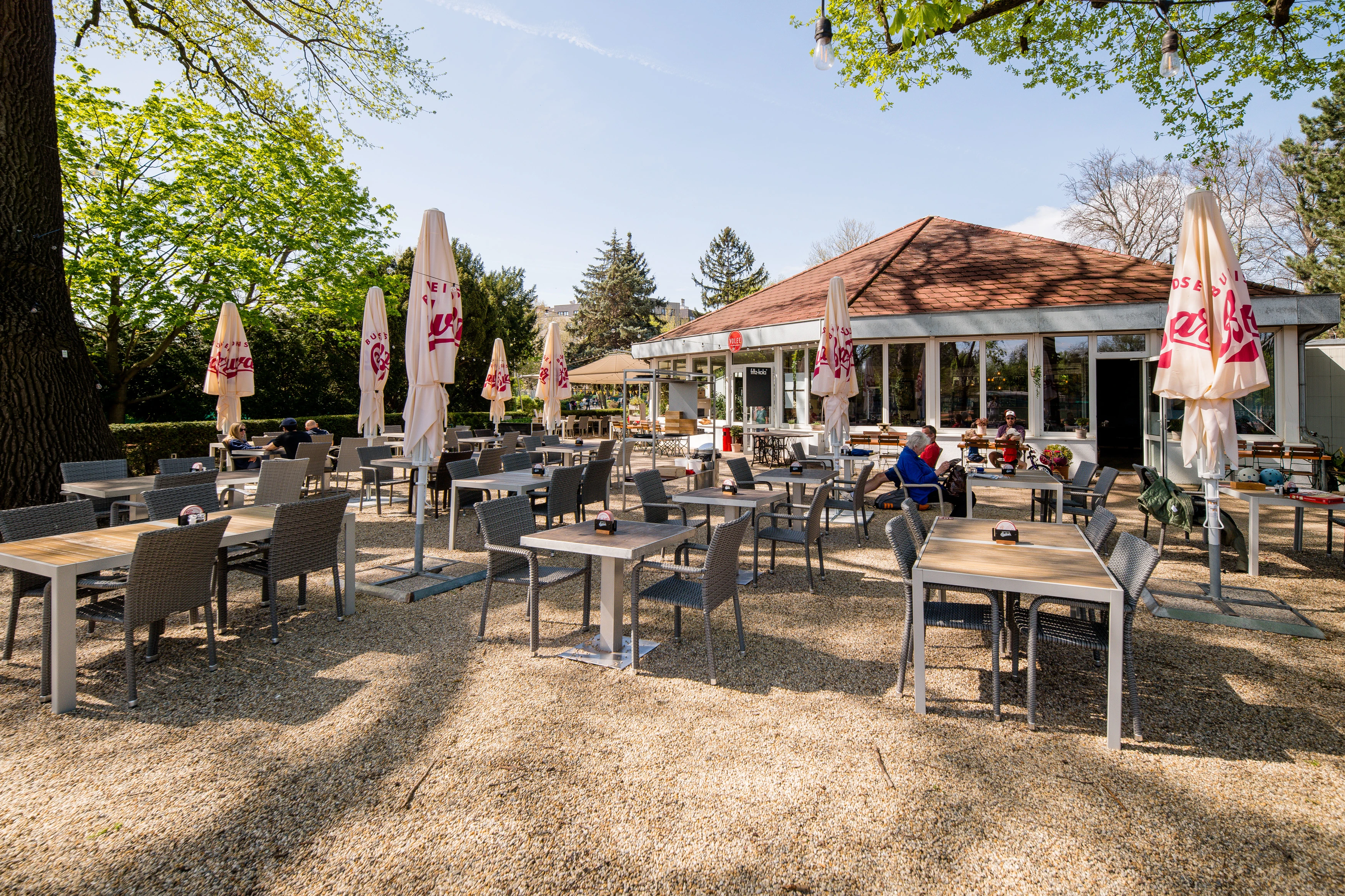 © Volee Einladender Biergarten mit Tischen, Stühlen und Sonnenschirmen unter Bäumen an einem sonnigen Tag.