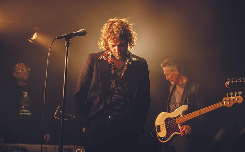 Voodoo Jürgens steht auf der Bühne mit einem Mikrofon vor sich, im Hintergrund sind zwei Bandmitglieder zu sehen