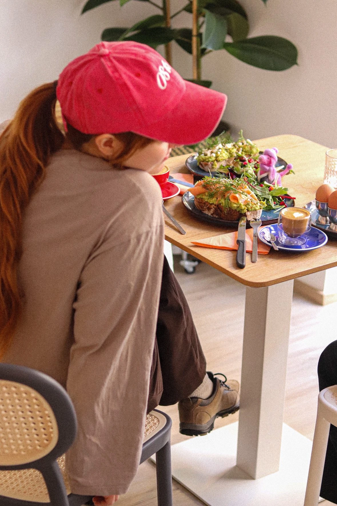 Frau mit roter Kappe genießt Avocado-Toast und Kaffee an einem hellen Tisch.