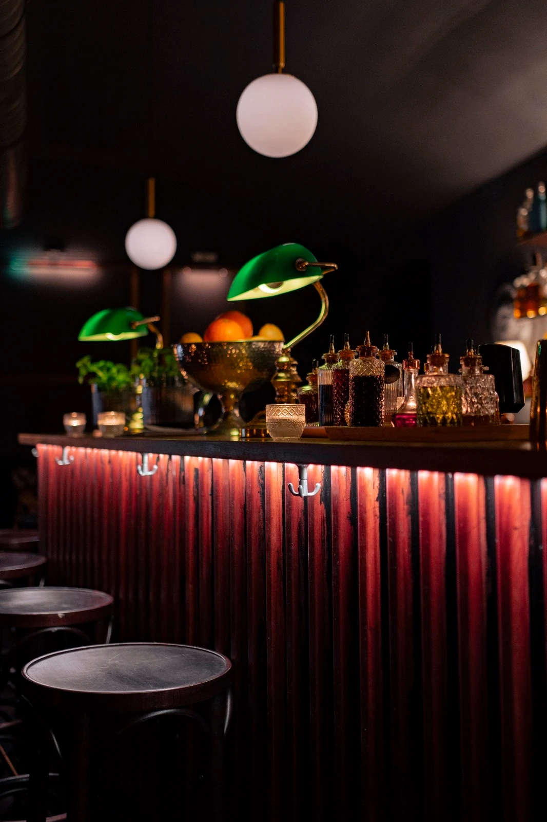 Atmosphärische Barszene mit Fokus auf Tresendetails: Barhocker, rote Theke, grüne Lampen, Dekanter und Obstschale.