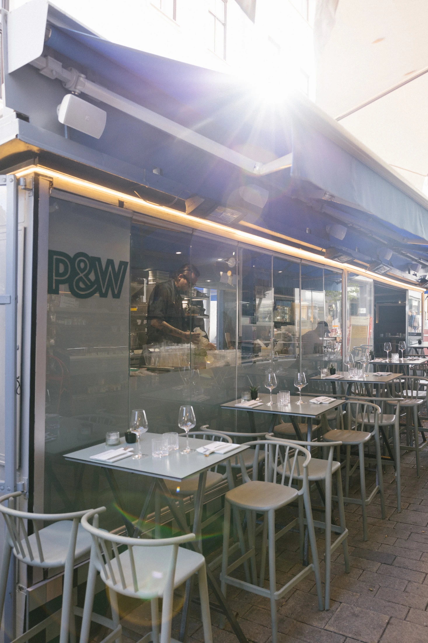 Außenansicht eines stilvollen Restaurants mit Sitzgelegenheiten im Freien und einem Blick in die Küche, in der ein Koch arbeitet.