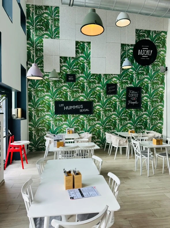 Restaurantinnenraum mit Blättertapete, weißen Möbeln und humorvollen Tafeln.