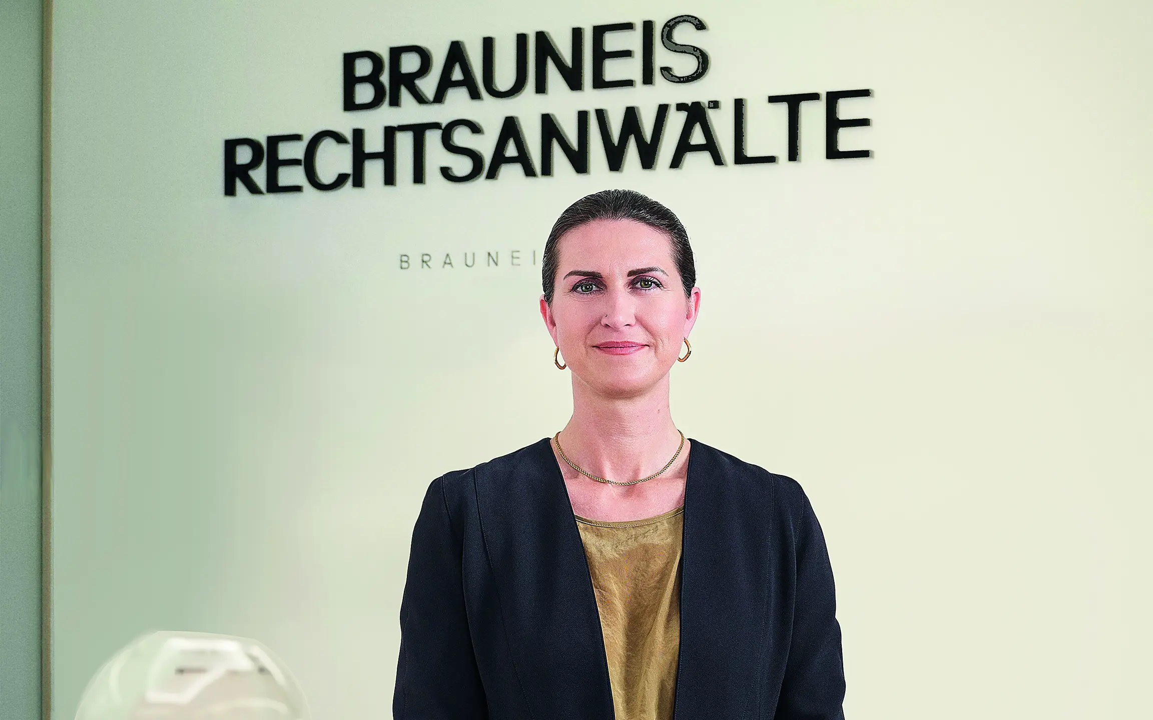 © Beigestellt Linda Poppenwimmer vor einer weißen Wand mit der Aufschrift „Brauneis Rechtsanwälte"