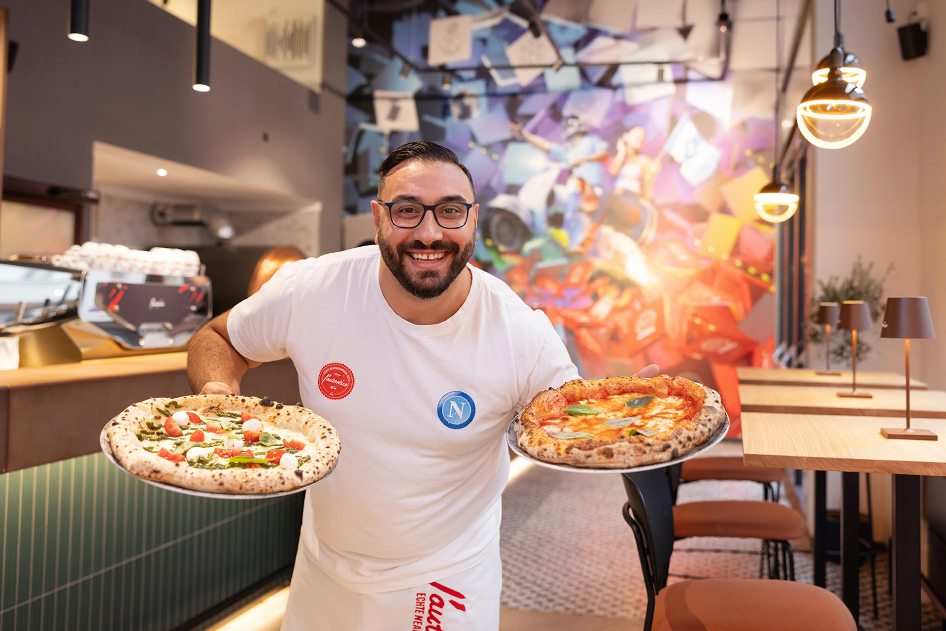 Lachender Mann mit Brille präsentiert zwei Pizzen im Restaurant.