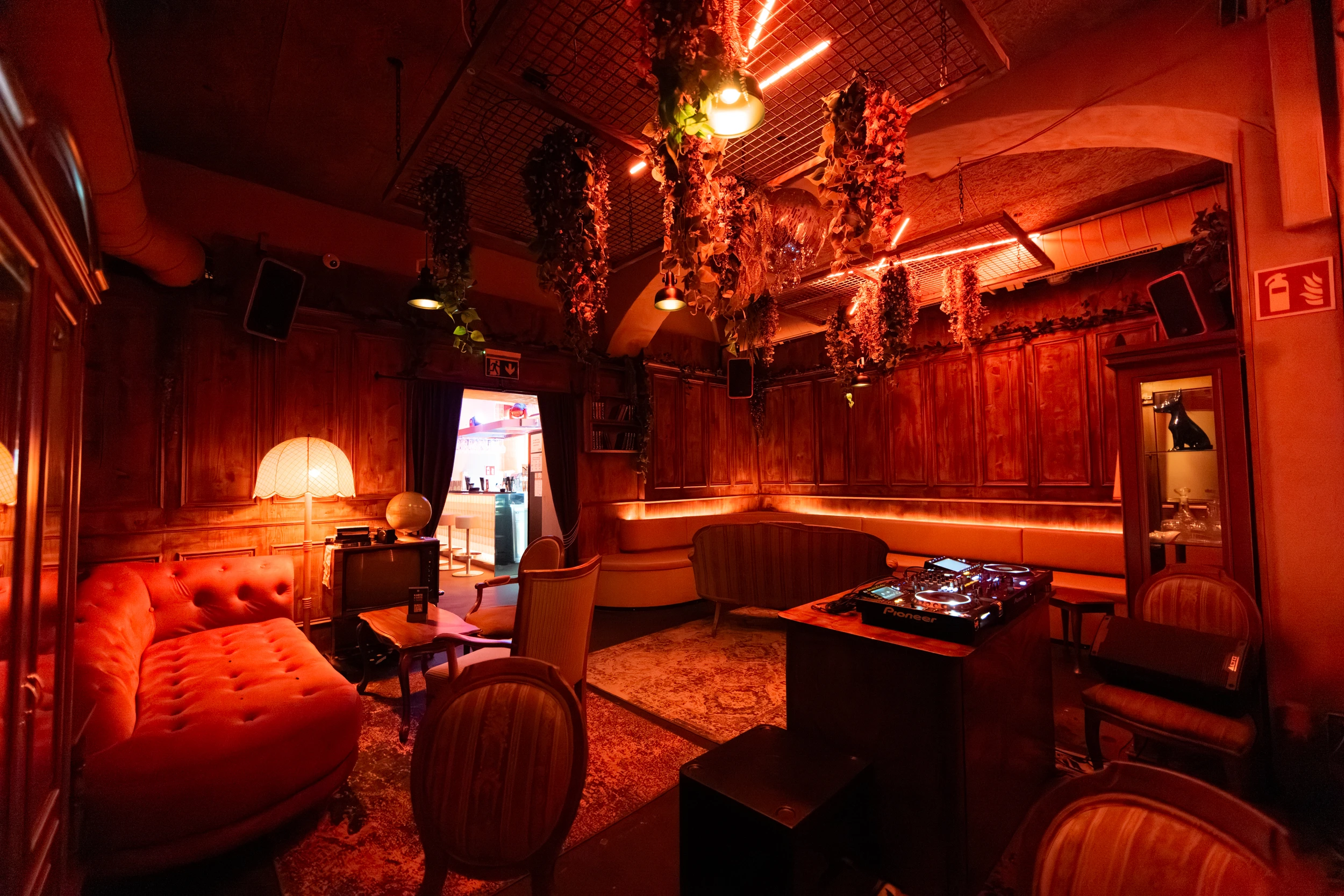 Bar Interieur mit rotem Samtsofa, DJ-Pult und Pflanzen an der Decke in gedämpfter roter Beleuchtung