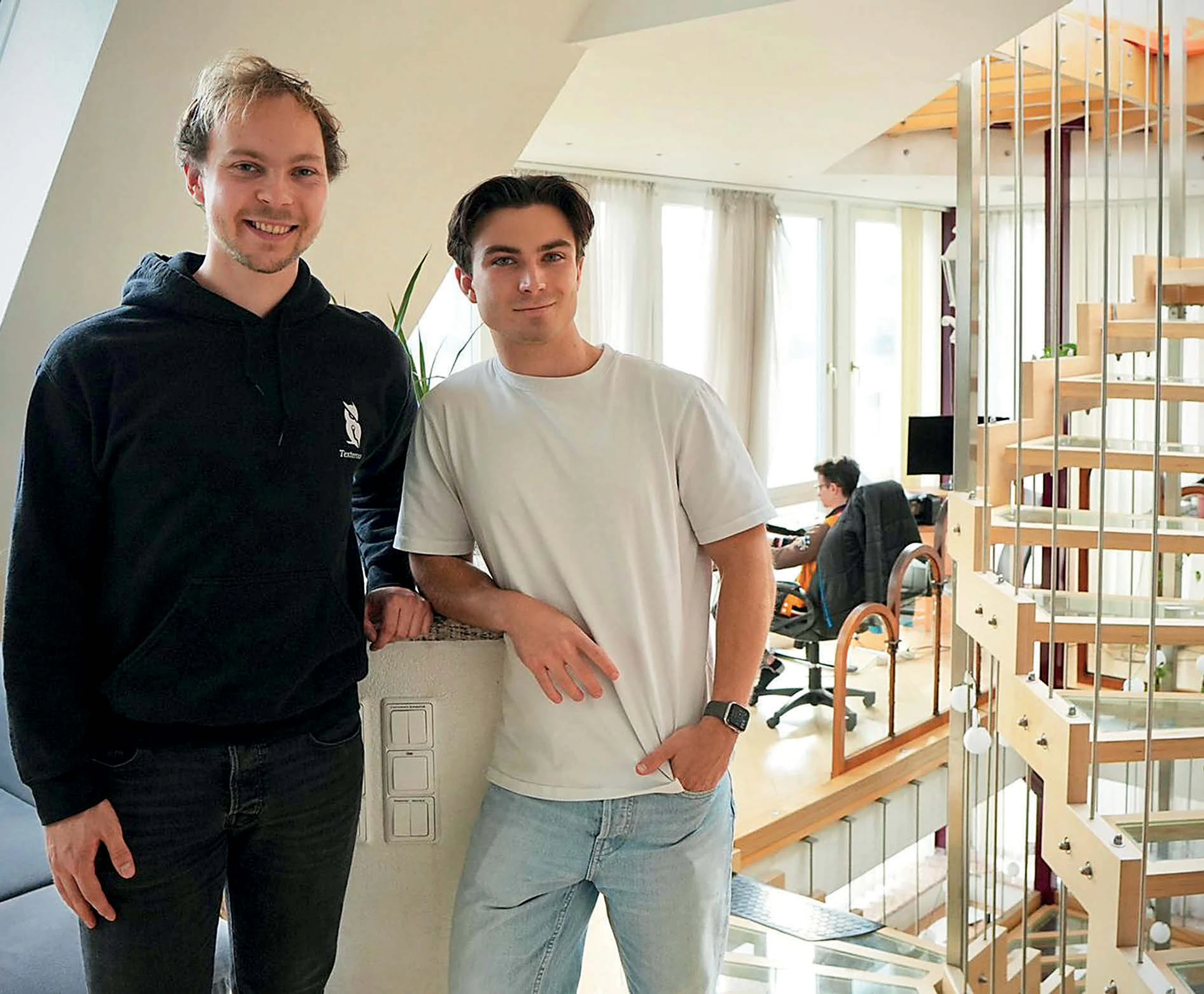 Zwei Männer in einem Wohnzimmer, Wendeltreppe rechts im Bild