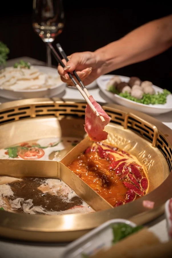 Hot Pot mit Fleisch und Essstäbchen. Das Fleisch wird in die Brühe getaucht. Im Hintergrund Speisen und Weinglas.