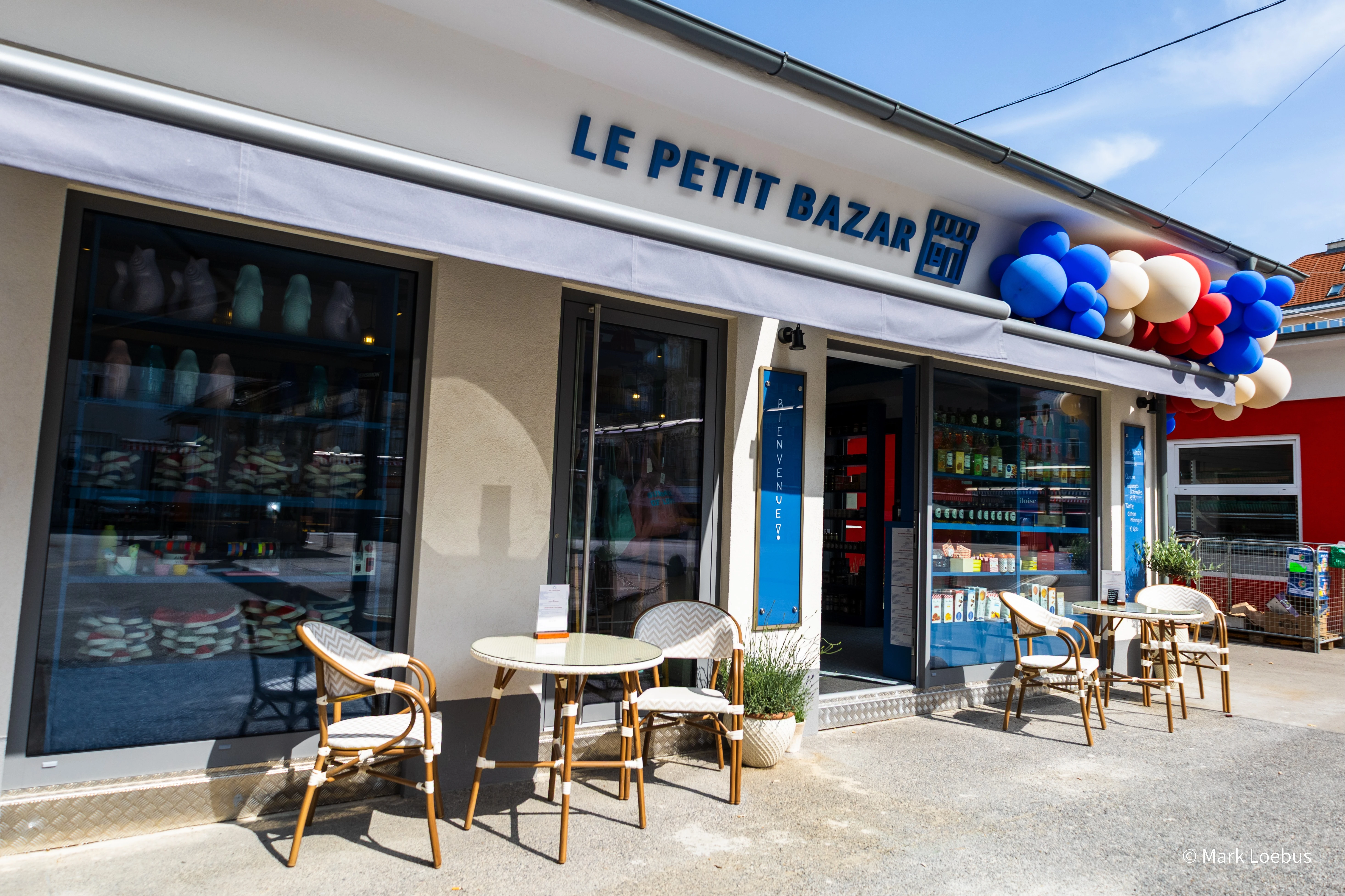 Fassade von Le Petit Bazar mit Tischen und Stühlen vor dem Laden, dekoriert mit Ballons.