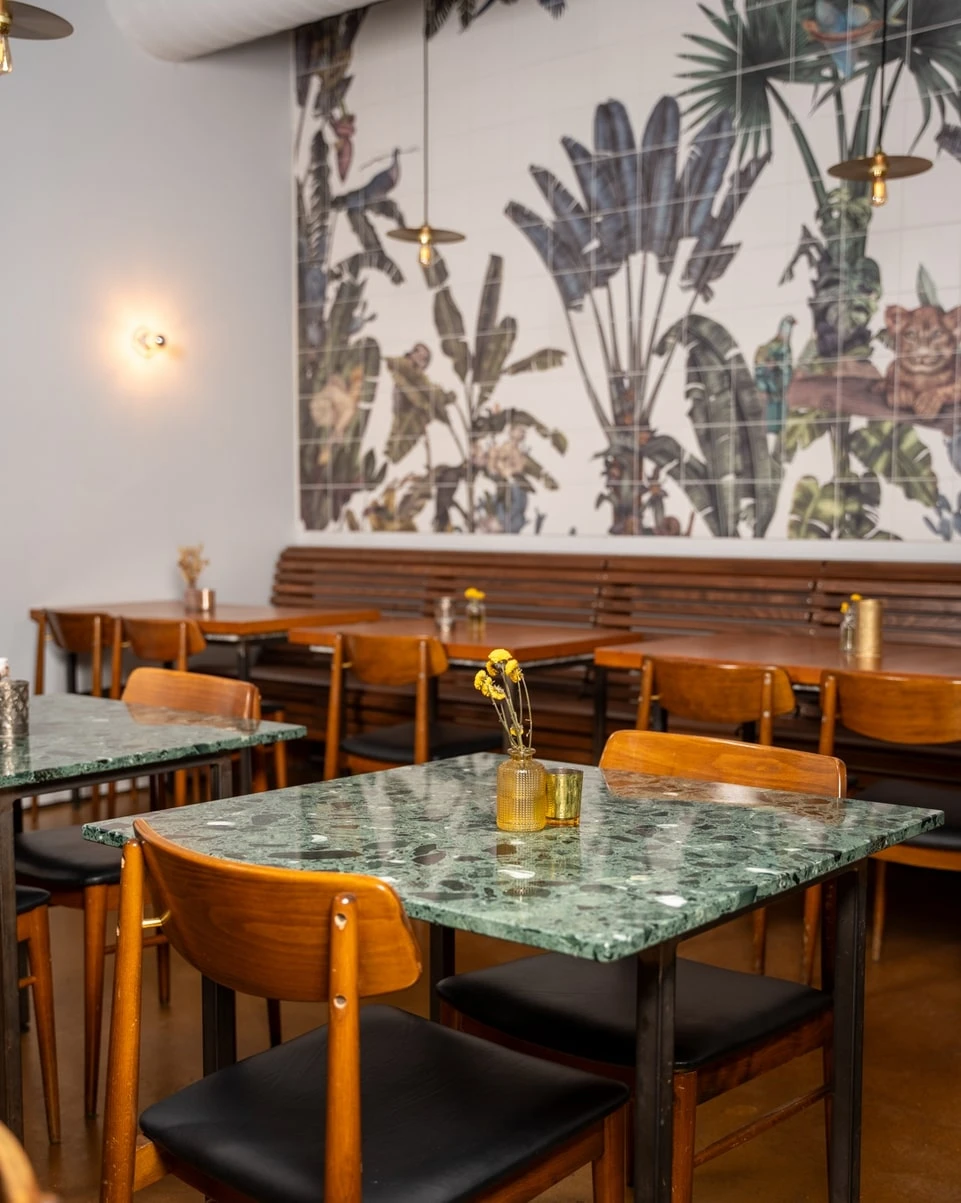 Restaurant mit Marmortischen, Holzstühlen und tropischem Wandbild.