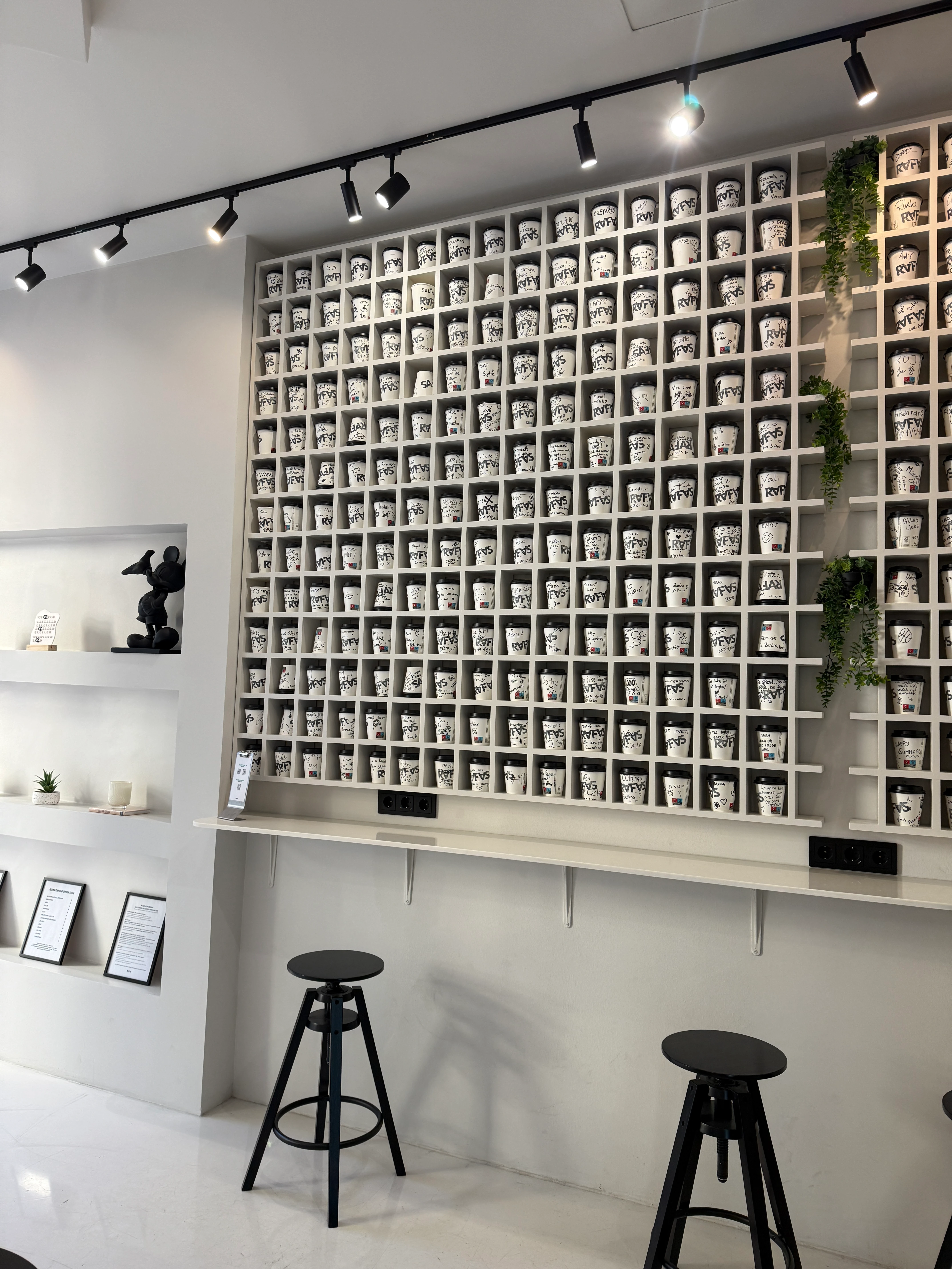 Café-Interieur mit Regalwand voller Kaffeebecher, schwarzen Hockern und dekorativen Elementen.