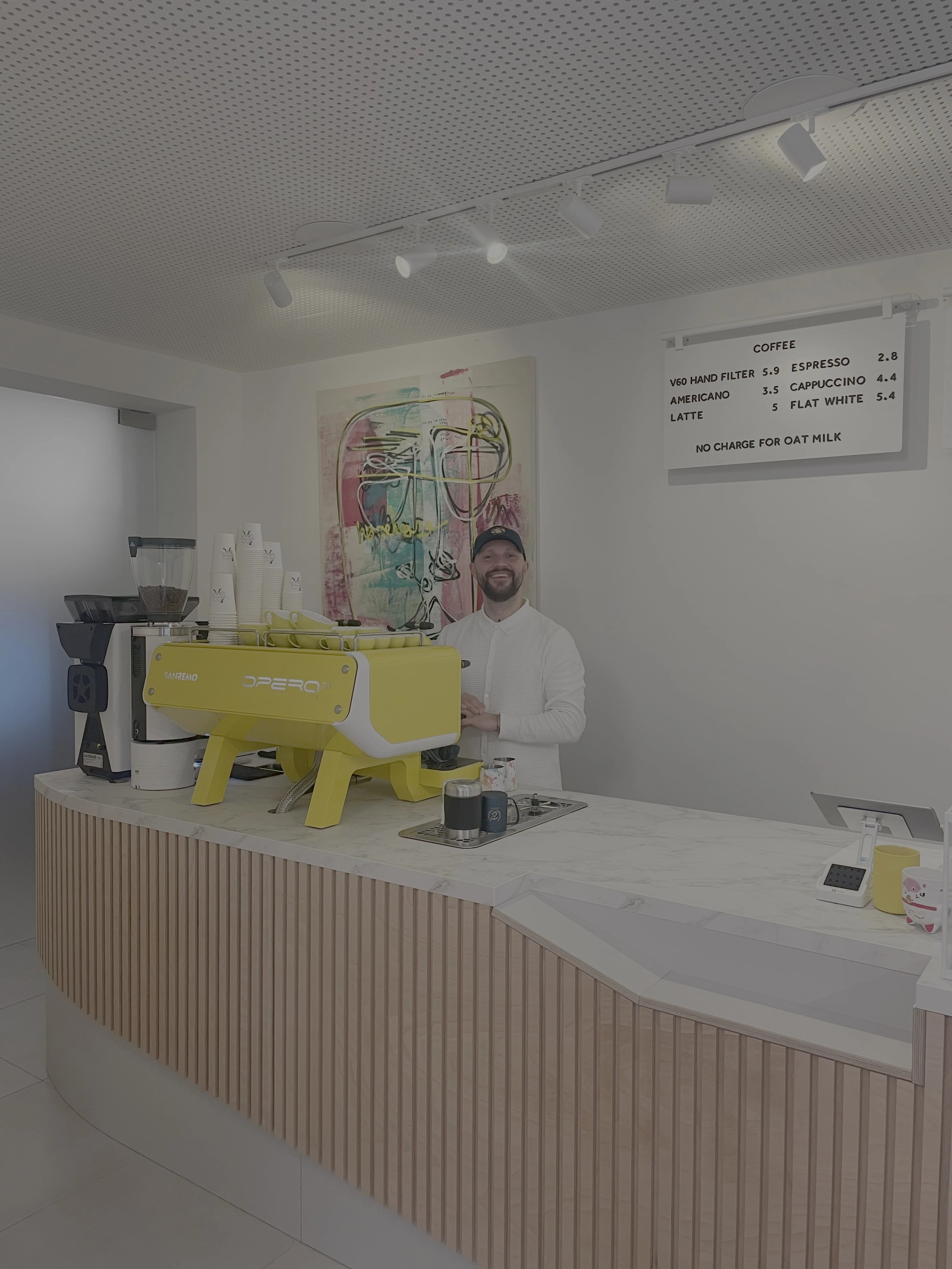 Moderne Kaffeebar mit Barista, Sanremo Kaffeemaschine und Kaffeekarte mit Preisen für Espresso, Cappuccino und mehr.