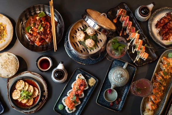 Vielfältige Auswahl an asiatischen Gerichten, kunstvoll auf einem dunklen Tisch präsentiert. Sushi, Dim Sum und weitere Spezialitäten.