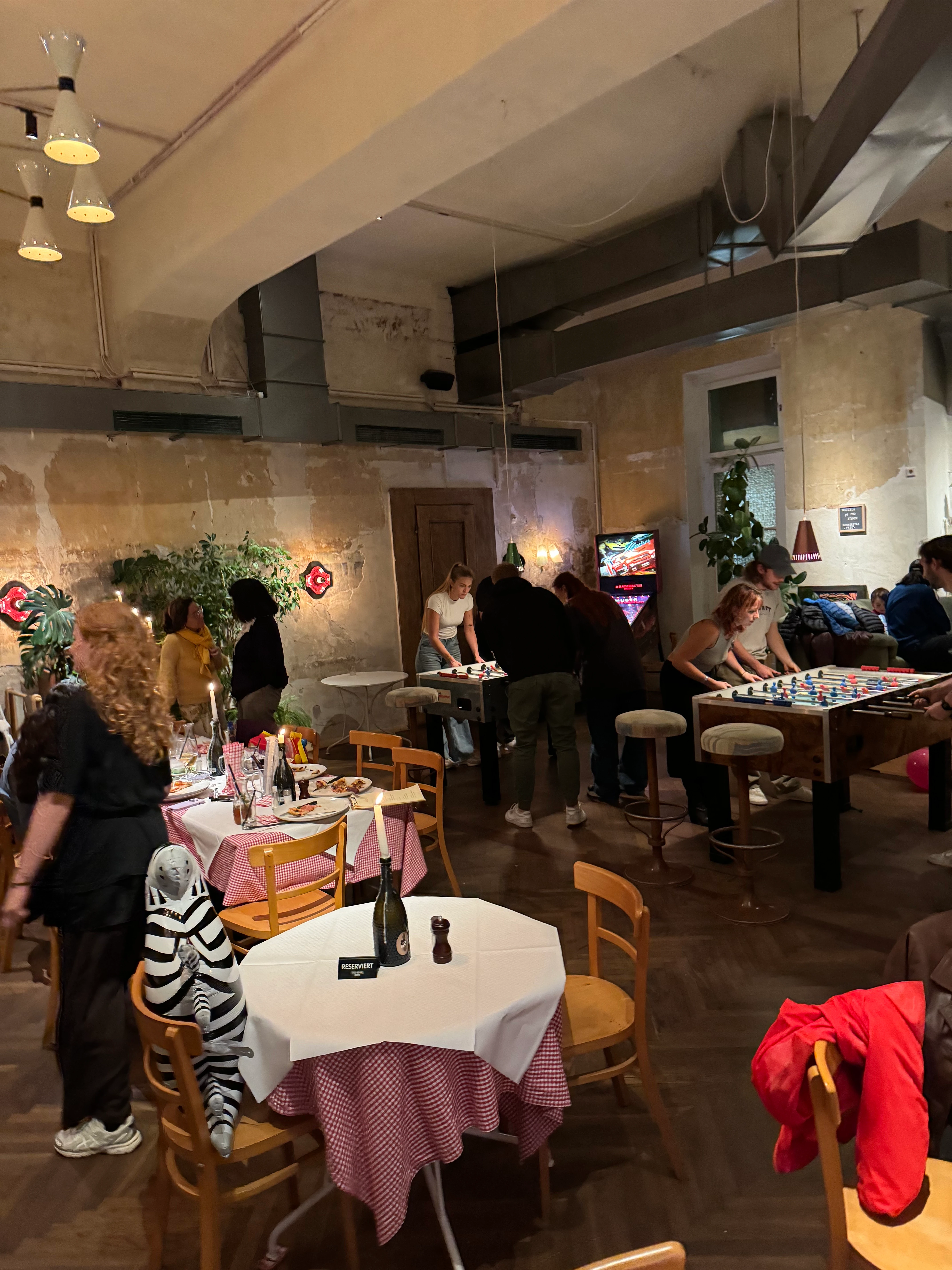 People enjoying dinner and playing foosball in a rustic restaurant or bar. The scene is filled with chatter, food, and a lively atmosphere.