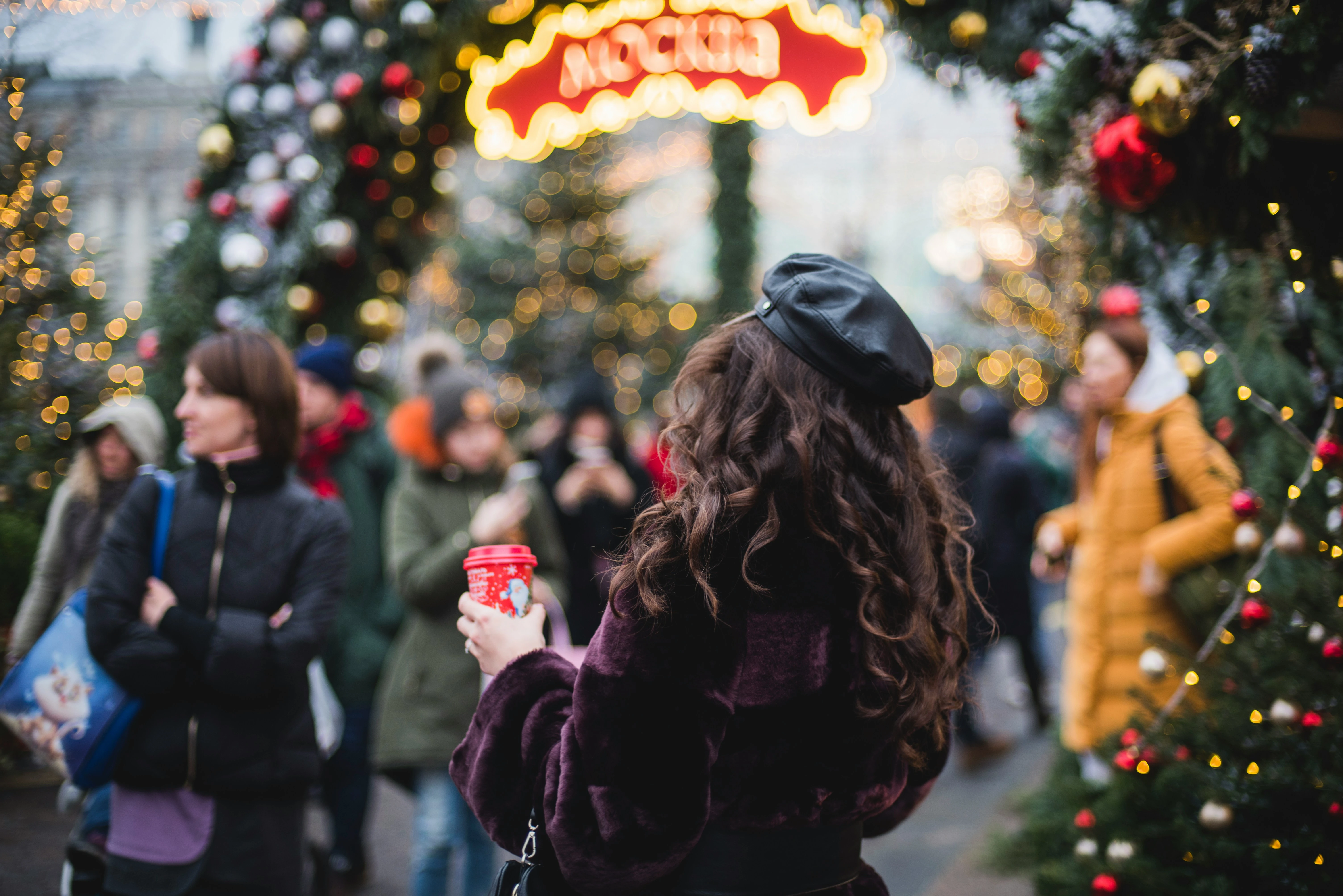 Frau mit Mütze und Becher auf einem stimmungsvollen Weihnachtsmarkt mit Lichtern und geschmückten Bäumen.