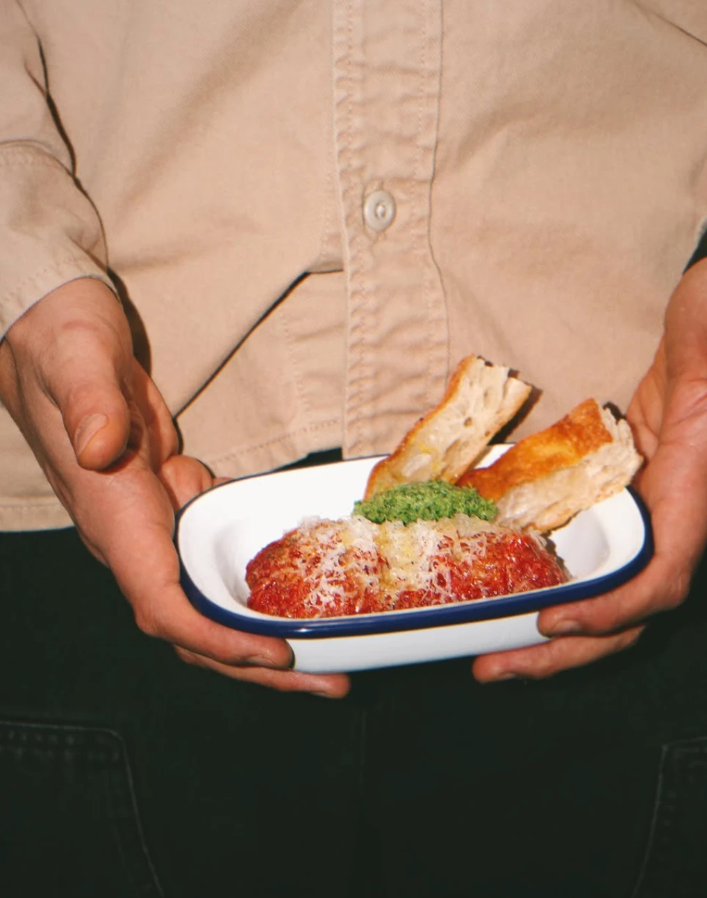 Person holding a plate of food featuring meat, tomato sauce, cheese, bread, and pesto.