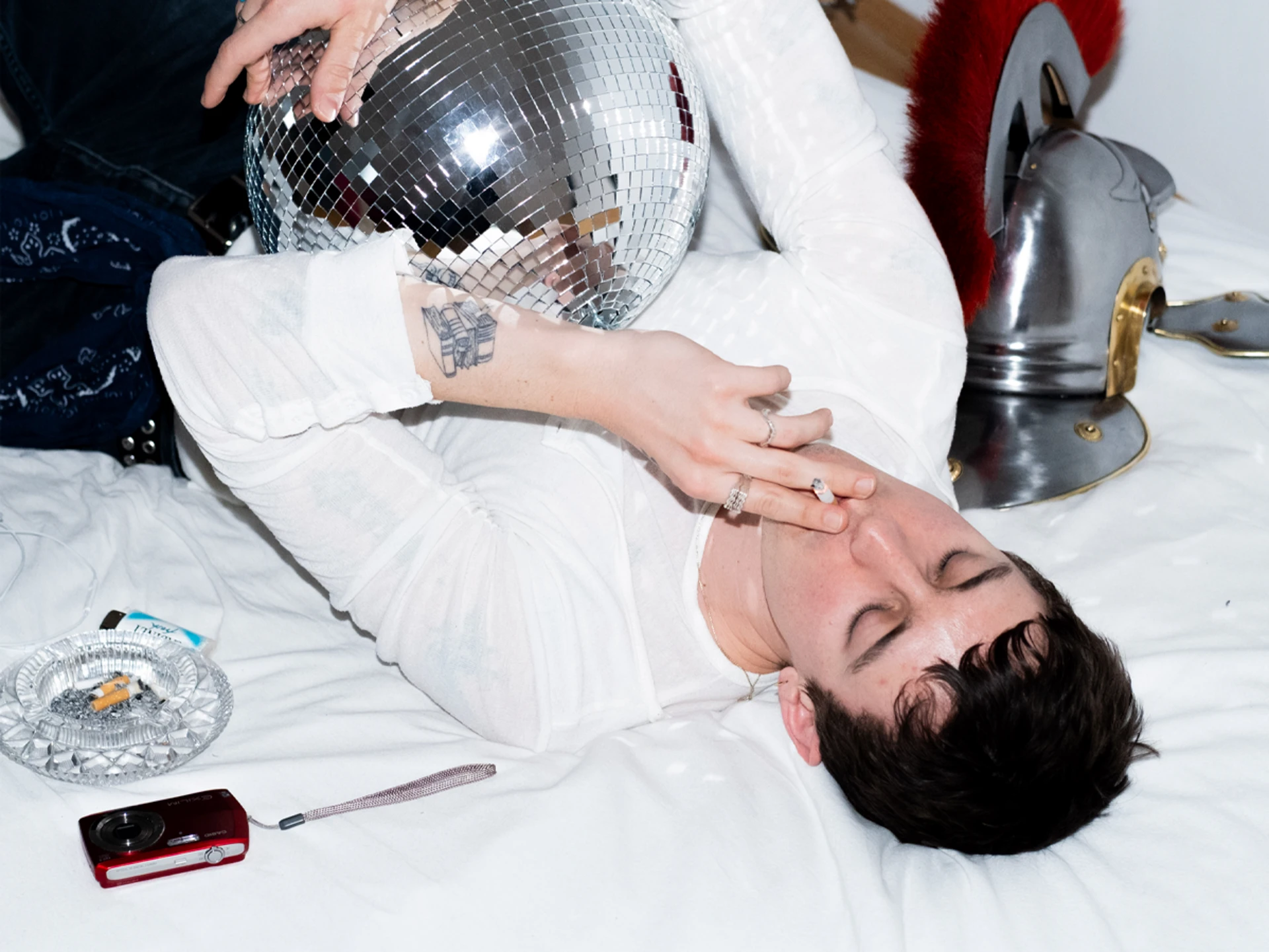 Mann liegt mit Discokugel, Zigarette, Kamera und römischem Helm auf dem Bett.