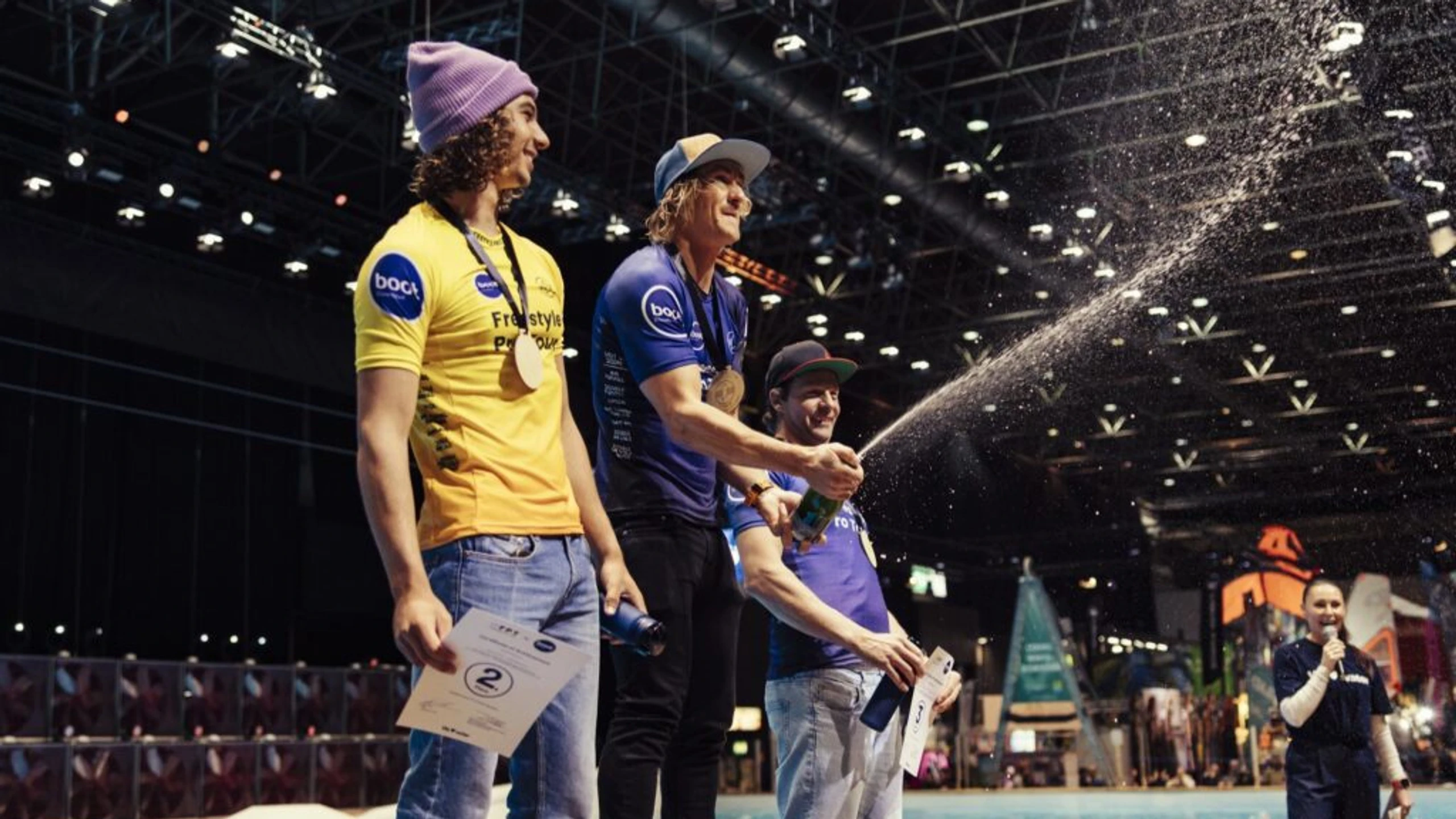 Drei Windsurf-Athleten feiern auf dem Podium am Beckenrand einer Indoor-Messehalle während der boot Düsseldorf 2026, umgeben von Publikum und Messeständen. Der Mann in der Mitte in blauem Shirt mit Medaille sprüht Champagner in die Luft, links steht ein Mann in gelbem Shirt mit Goldmedaille und purpurner Beanie, rechts einer in violettem Shirt mit Silbermedaille; Sektfontäne im Vordergrund, blaues Wasser und beleuchtete Halle im Hintergrund.