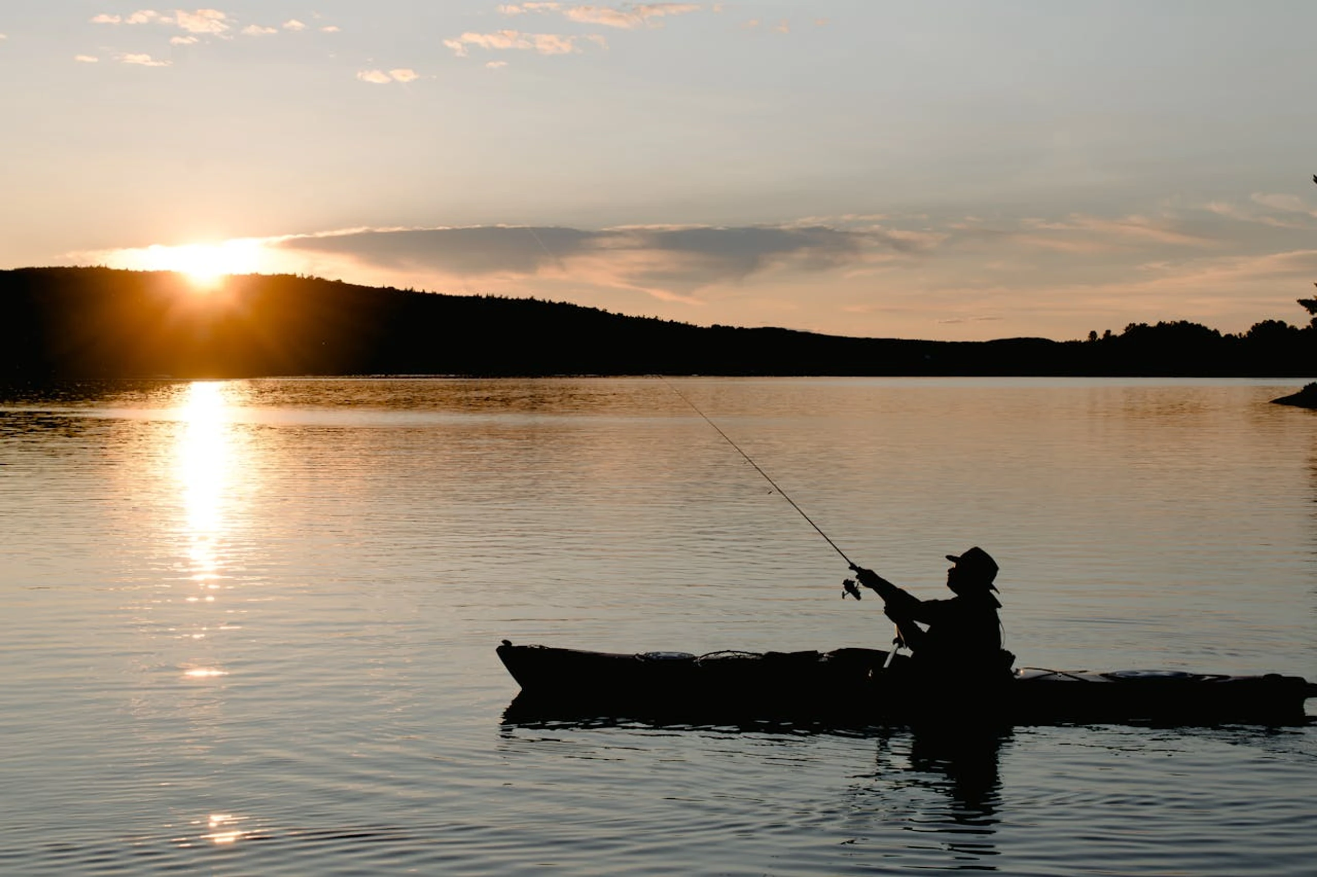 Silhouette eines Anglers in einem Kanu bei Sonnenuntergang auf einem See: Der Hobbyfischer wirft die Rute aus, im Hintergrund spiegelt sich die Sonne auf dem Wasser mit bewaldeten Hügeln. Symbol für entspannte Freizeitangeln – wie es bald mit der EU-App RecFishing einfacher gemeldet wird.