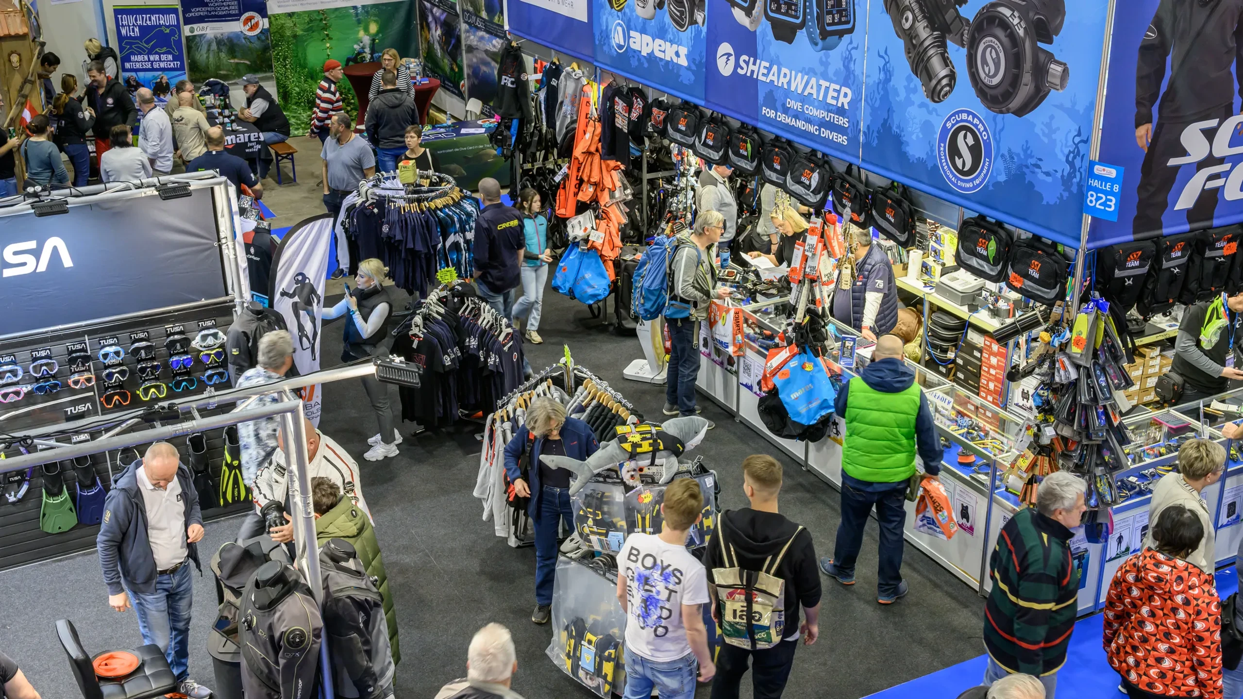 Lebendige Messehalle der BOOT TULLN mit belebter Tauch-Ausstellerzone: Besucher in Alltagskleidung schlendern zwischen Ständen mit Neoprenanzügen, Flossen, Computern und Ausrüstung. Große Banner von Shearwater, Scubapro und Subsanea rahmen Regale mit Masken und Computern ein – dynamische Atmosphäre voller Entdeckungen.