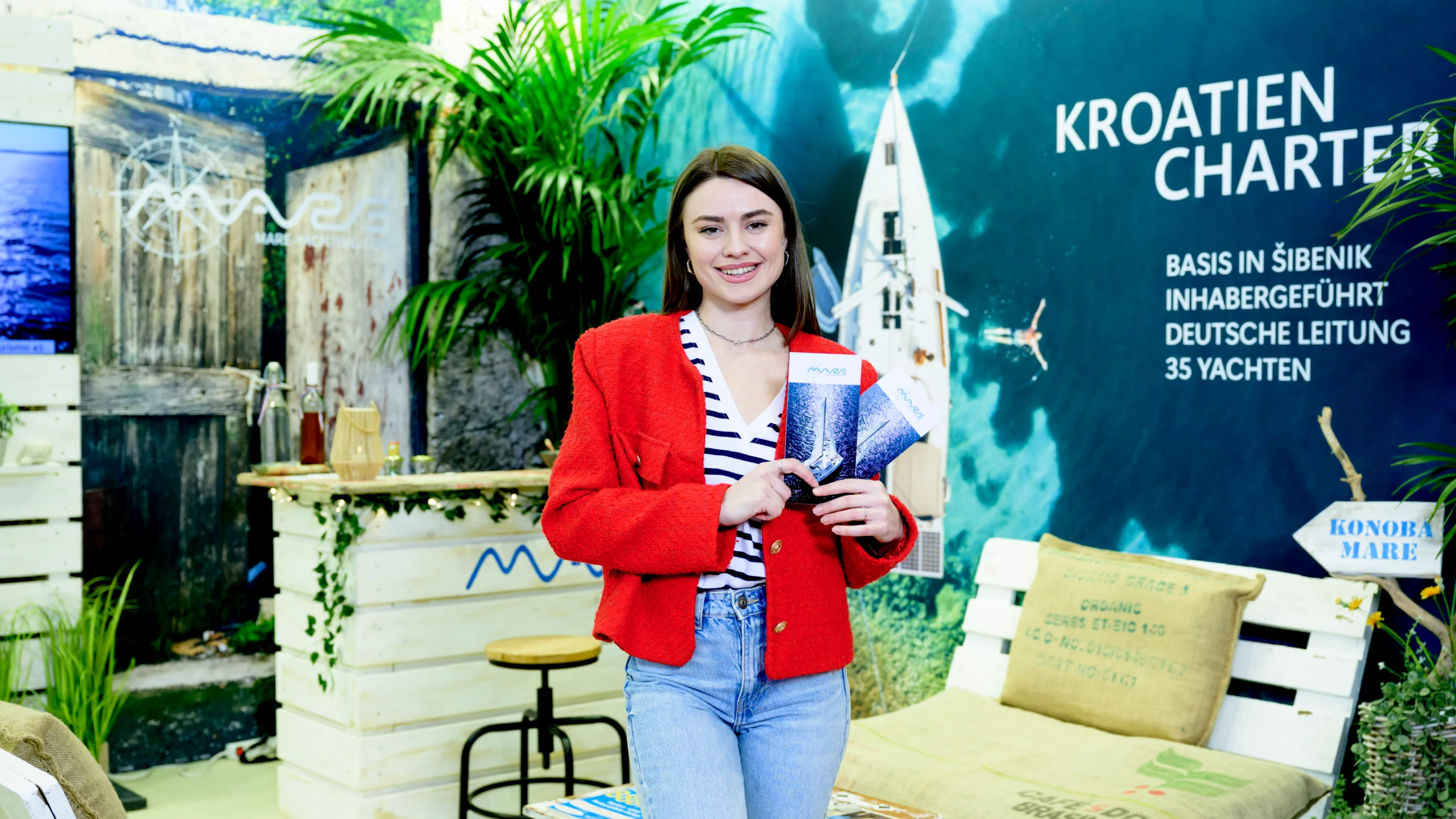Junge Frau in roter Jacke hält Prospekte an einem Messestand „Kroatien Charter“; im Hintergrund maritime Deko mit Palmen, Theke und Liegefläche