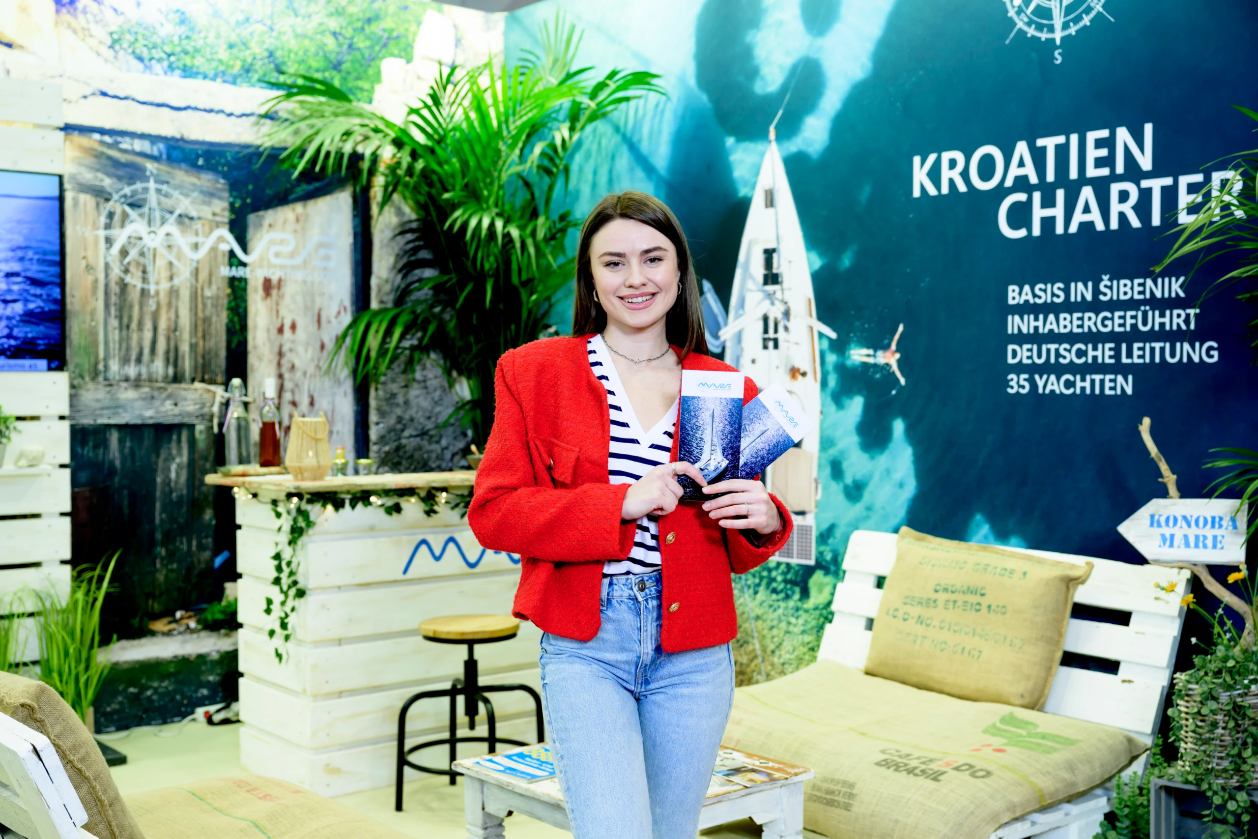 Junge Frau in roter Jacke hält Prospekte an einem Messestand „Kroatien Charter“; im Hintergrund maritime Deko mit Palmen, Theke und Liegefläche
