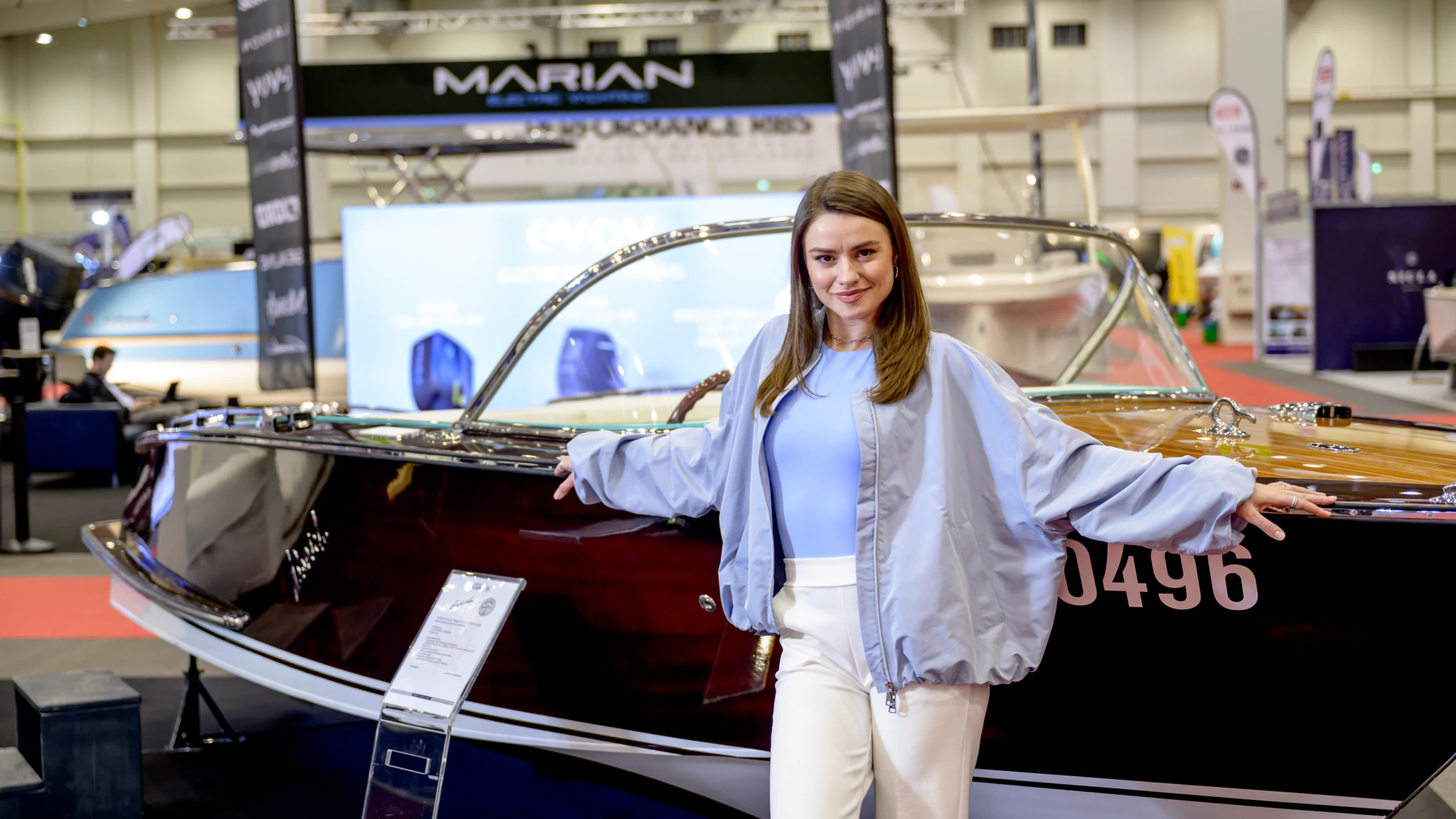 Junge Frau lehnt in einer Messehalle an einem glänzenden Motorboot mit Holzdeck; im Hintergrund weitere Bootsaussteller und Messestände.