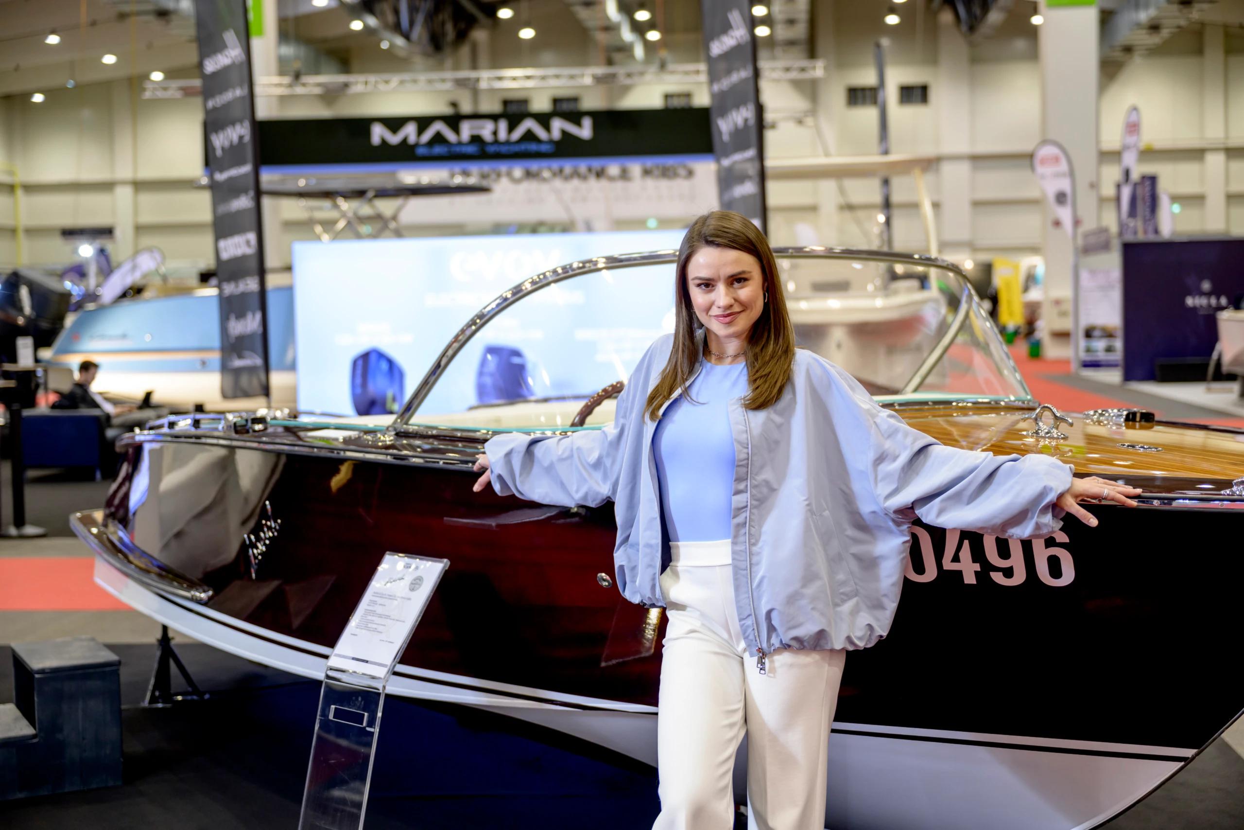 Junge Frau lehnt in einer Messehalle an einem glänzenden Motorboot mit Holzdeck; im Hintergrund weitere Bootsaussteller und Messestände.