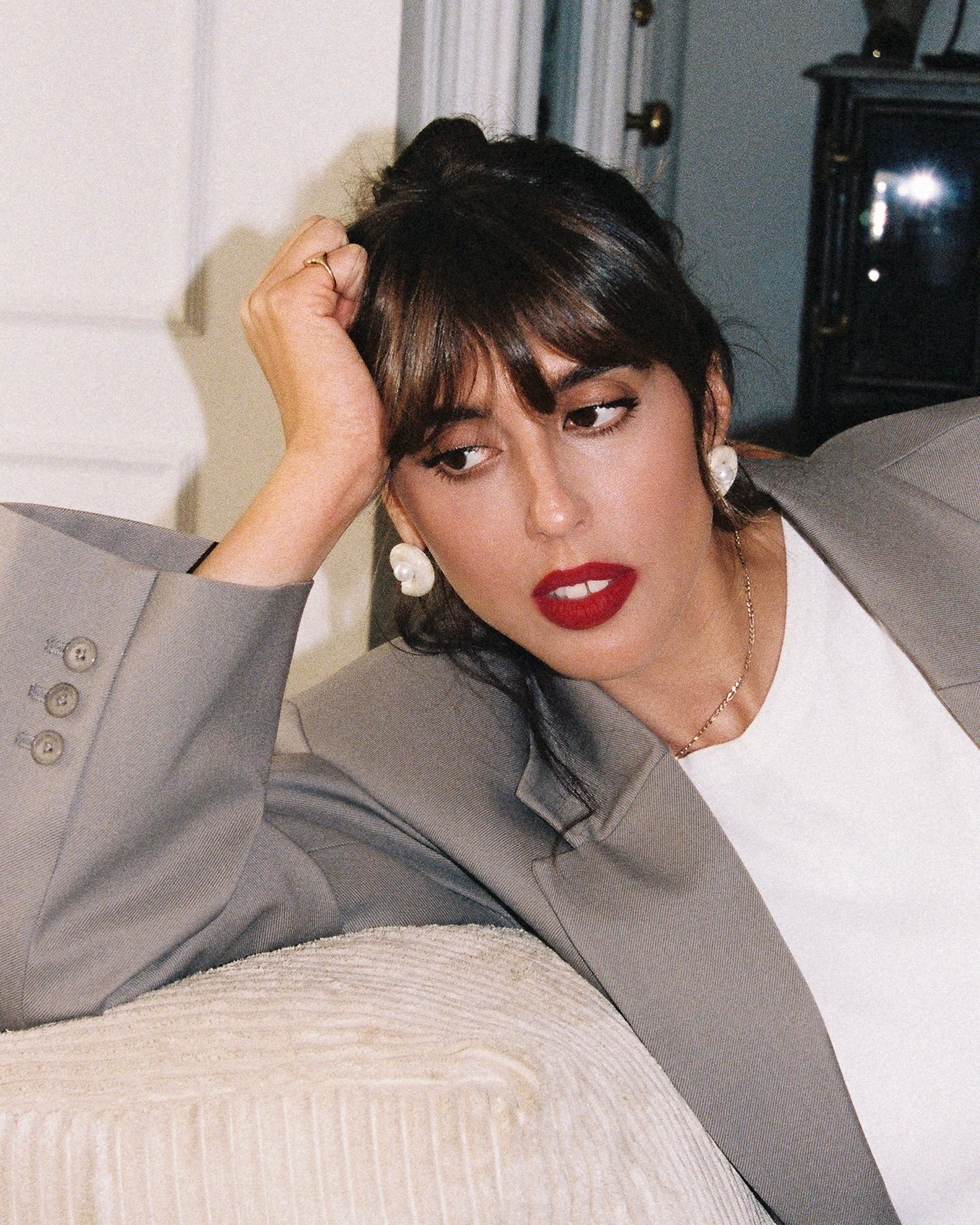 Violette Serrat lohnend auf einer Couch in grauem Blazer, weißem Shirt und rotem Lippenstift