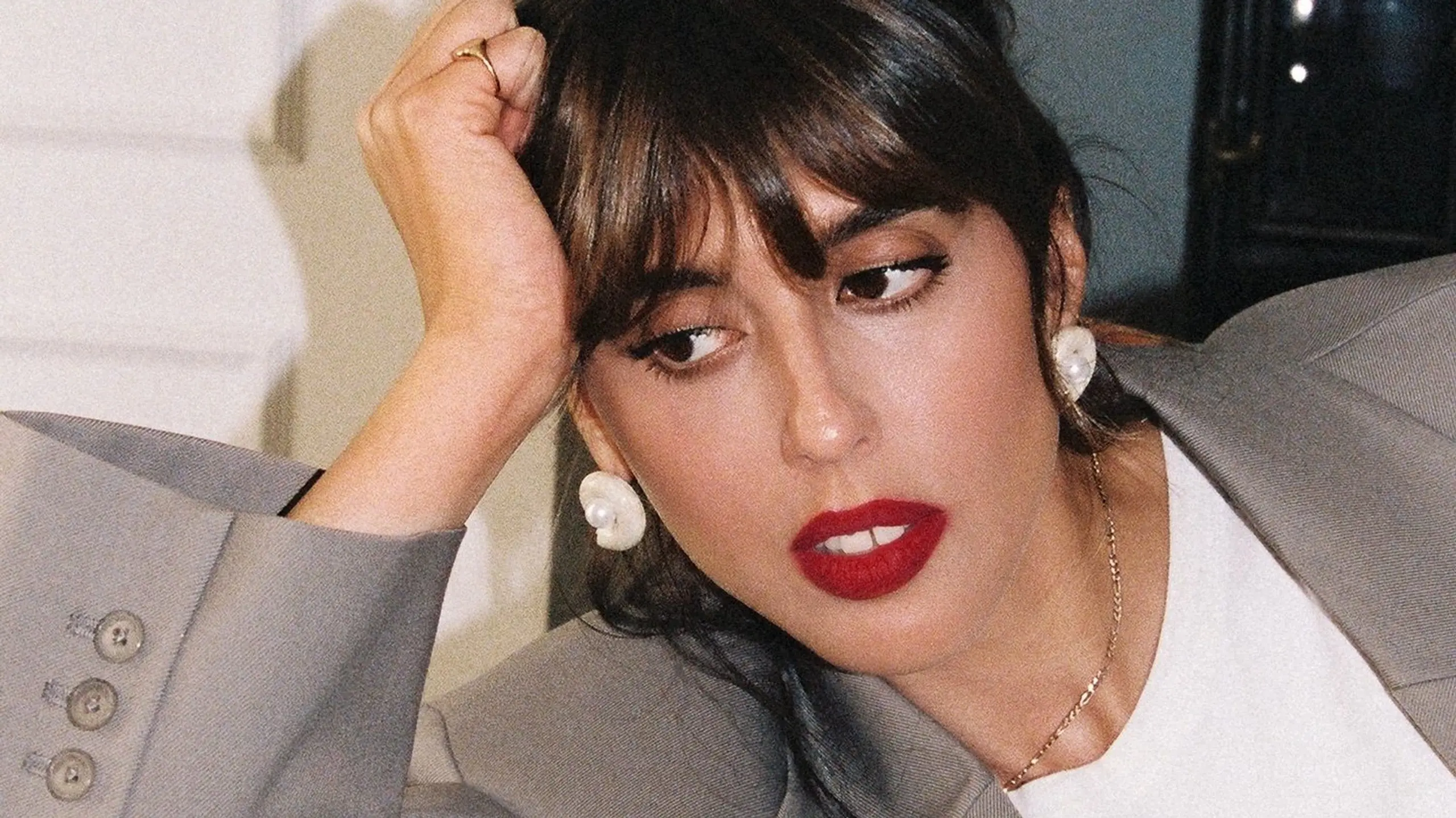 Violette Serrat lohnend auf einer Couch in grauem Blazer, weißem Shirt und rotem Lippenstift