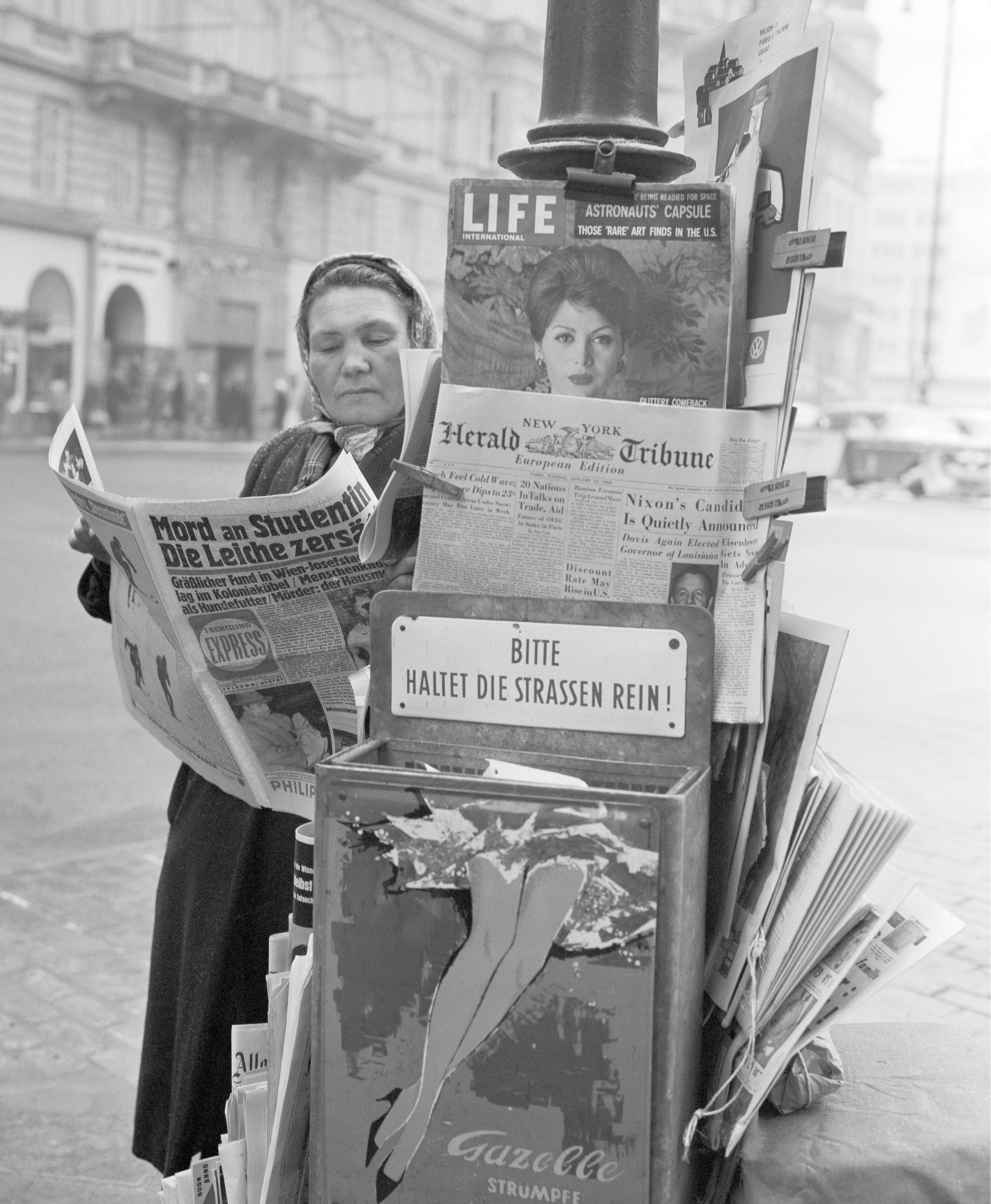 Eine Zeitungsverkäuferin steht an der Ecke Kärntnerstraße und Philharmonikerstraße in Wien um 1960. Im Hintergrund ist das Palais Todesco zu sehen. Die Szene fängt das Straßenbild und den Alltag Wiens in den 1960er Jahren ein.