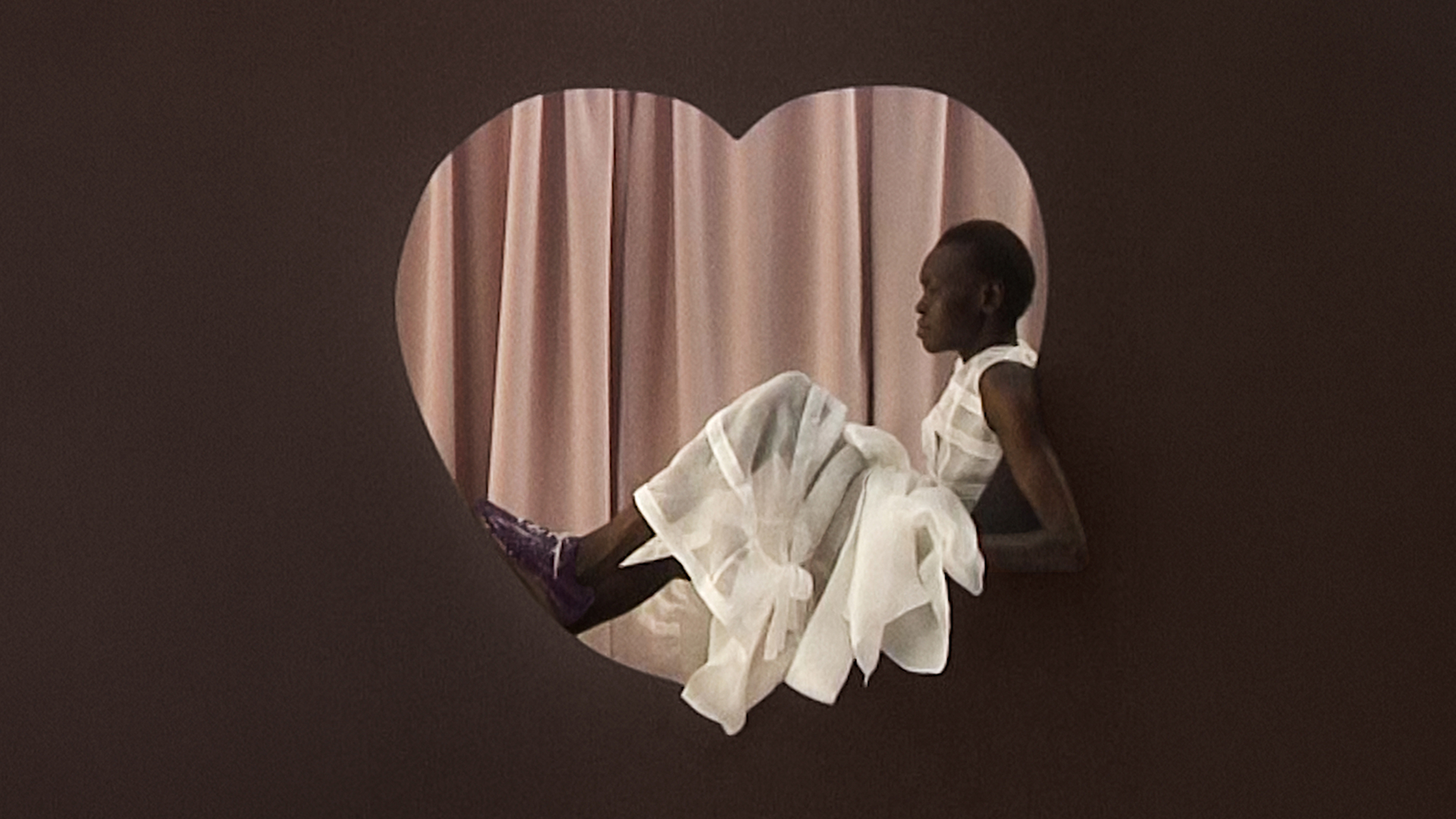 Alek Wek sitzt in herform mit weißem Kleid und lila Lackleder Schuhen