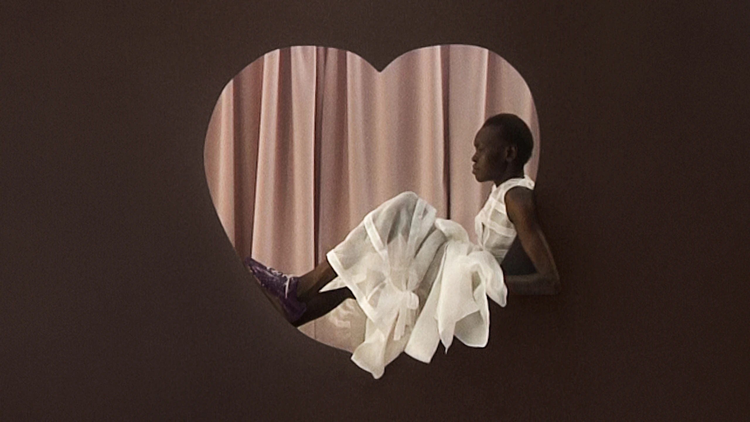 Alek Wek sitzt in herform mit weißem Kleid und lila Lackleder Schuhen