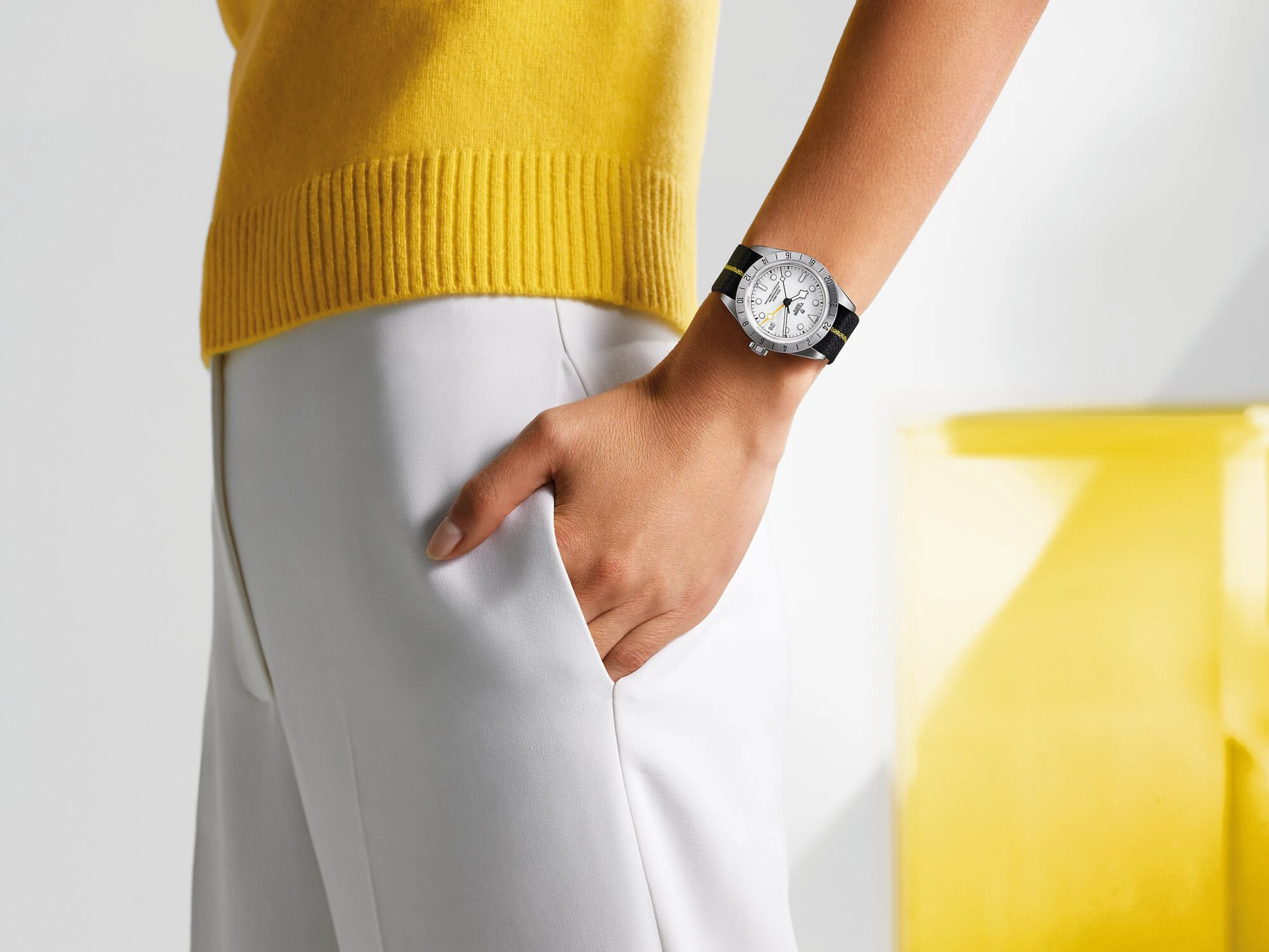 man sieht den Arm einer Person, die eine Uhr trägt. Sie trägt eine weiße Hose und einen gelben Pullover.